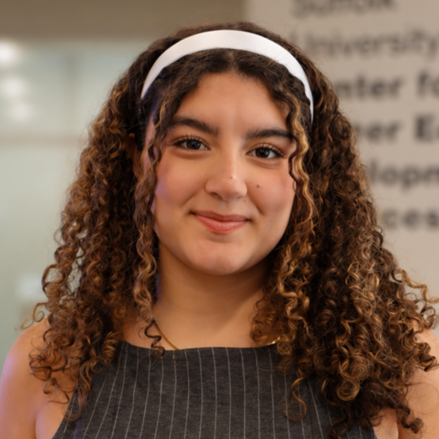 A woman with curly brown hair and light skin, wearing a dark top, looking directly at the camera against a gray background.