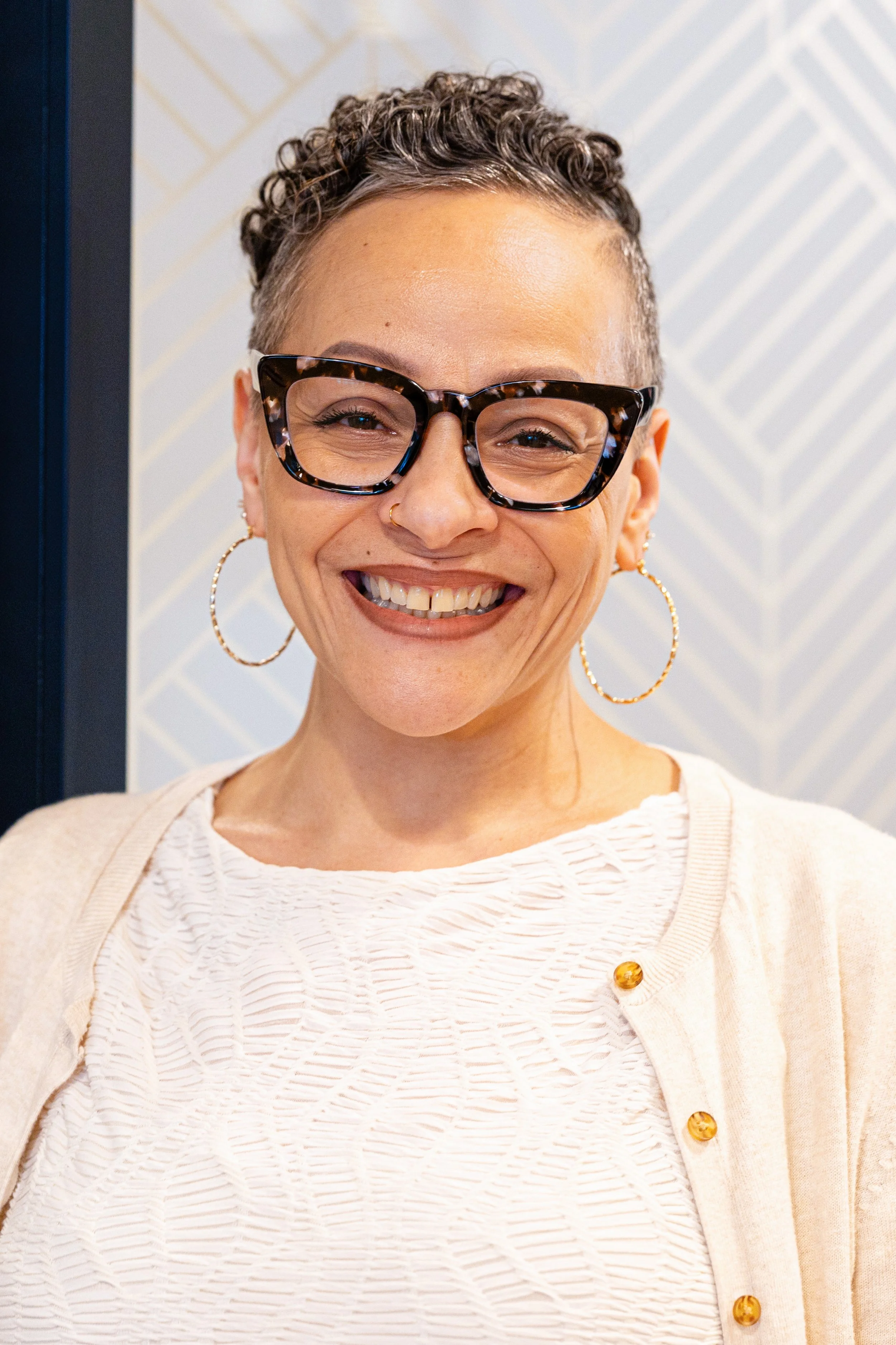 A woman with short, curly hair wearing large tortoiseshell glasses, gold hoop earrings, a nose ring, and a cream-colored cardigan over a textured white top, smiling happily.