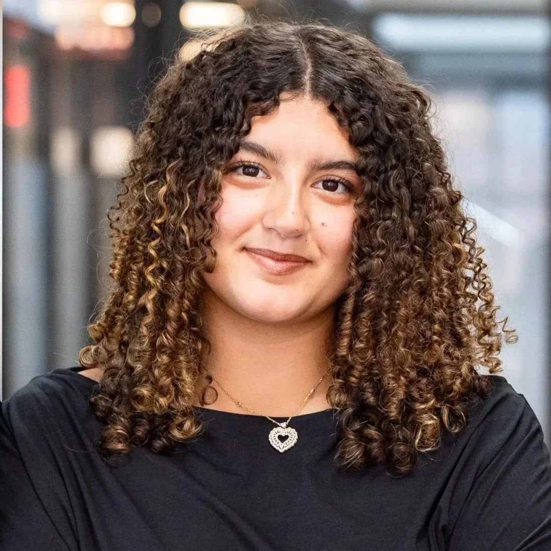 A woman with curly brown hair and light skin, wearing a dark top, looking directly at the camera against a gray background.