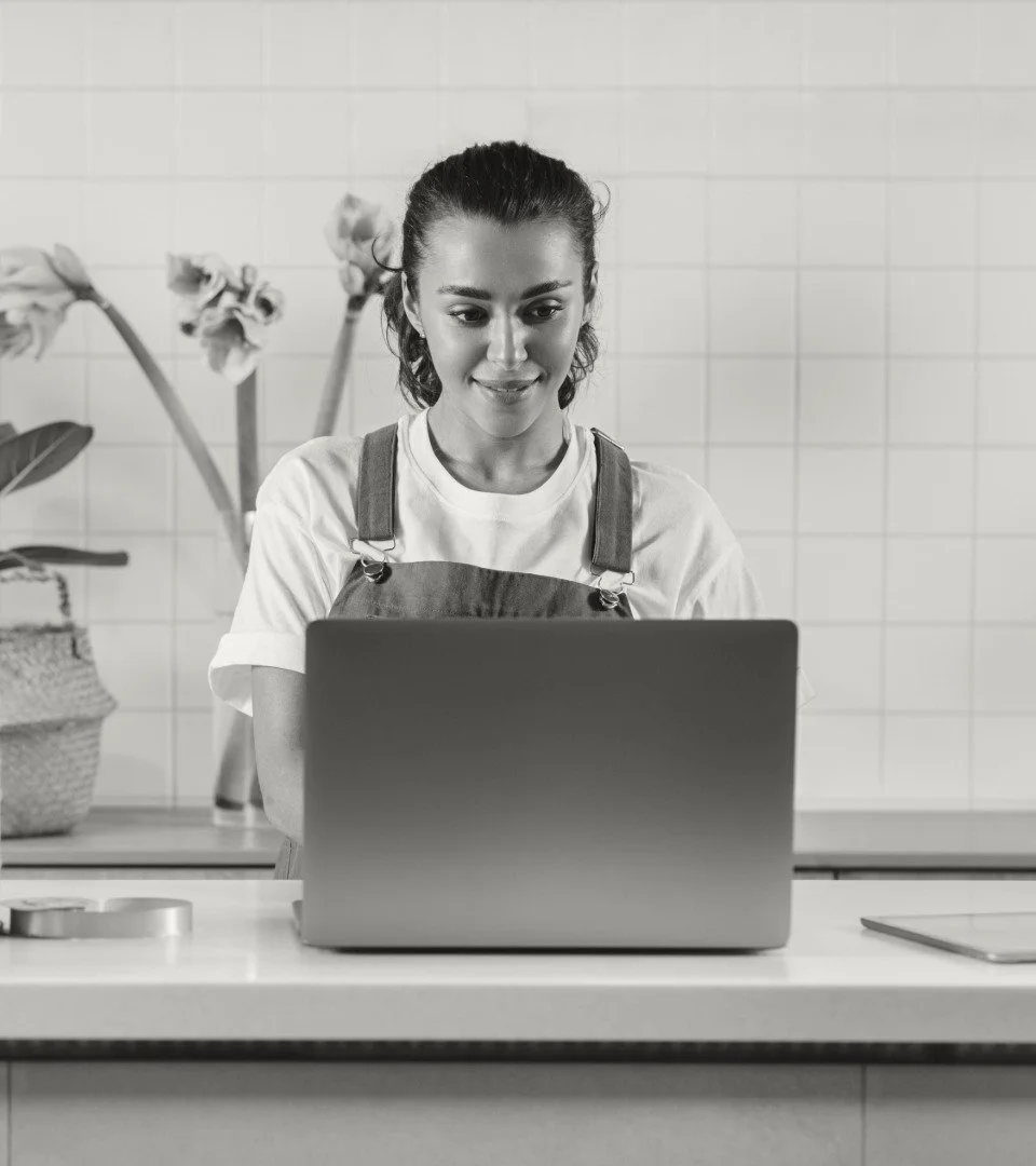 A woman in a white T-shirt and apron looks at her laptop screen in a kitchen.