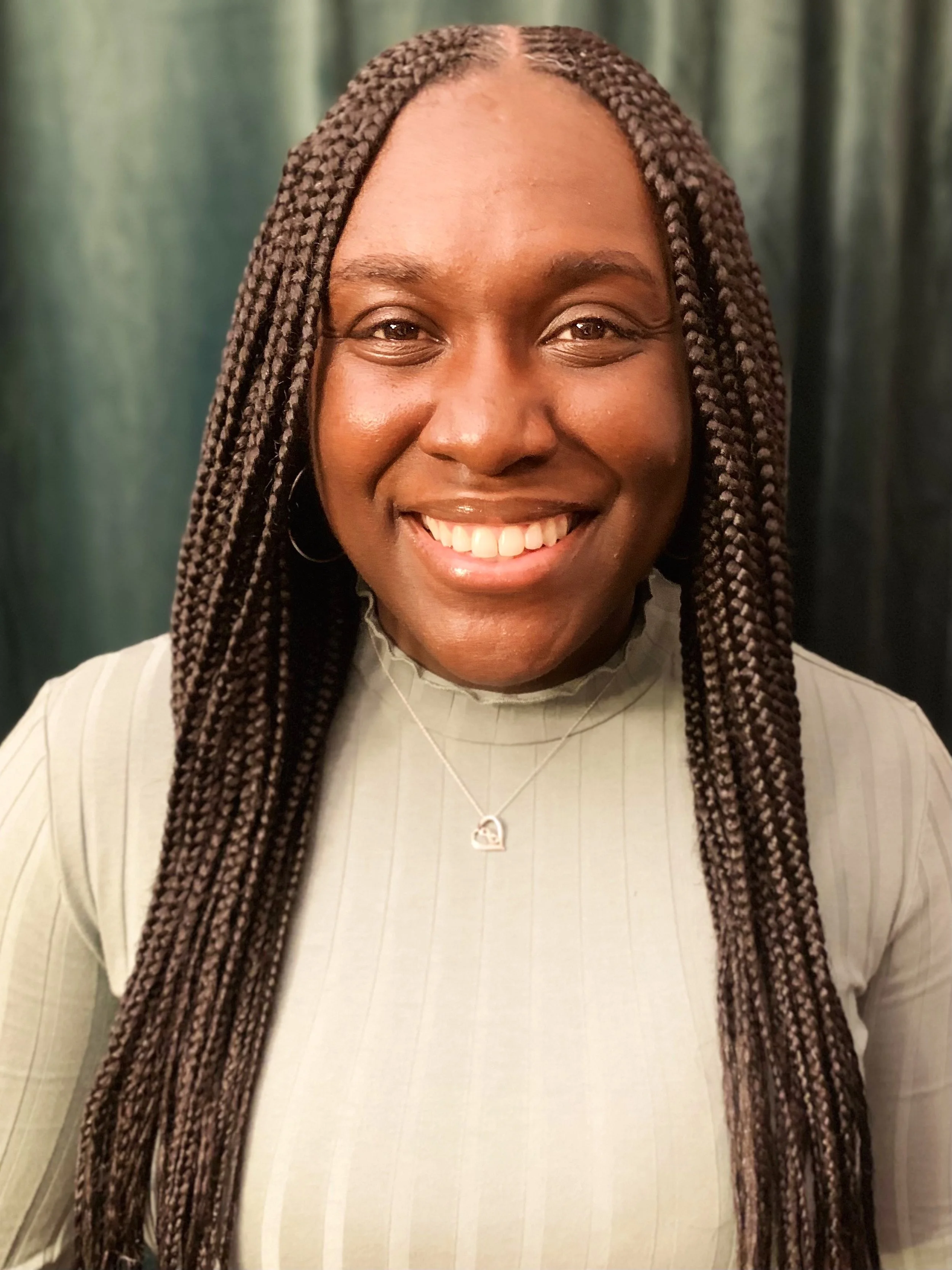 Close-up of a smiling woman with brown braids wearing a light-colored top and a necklace, with a dark green curtain background.