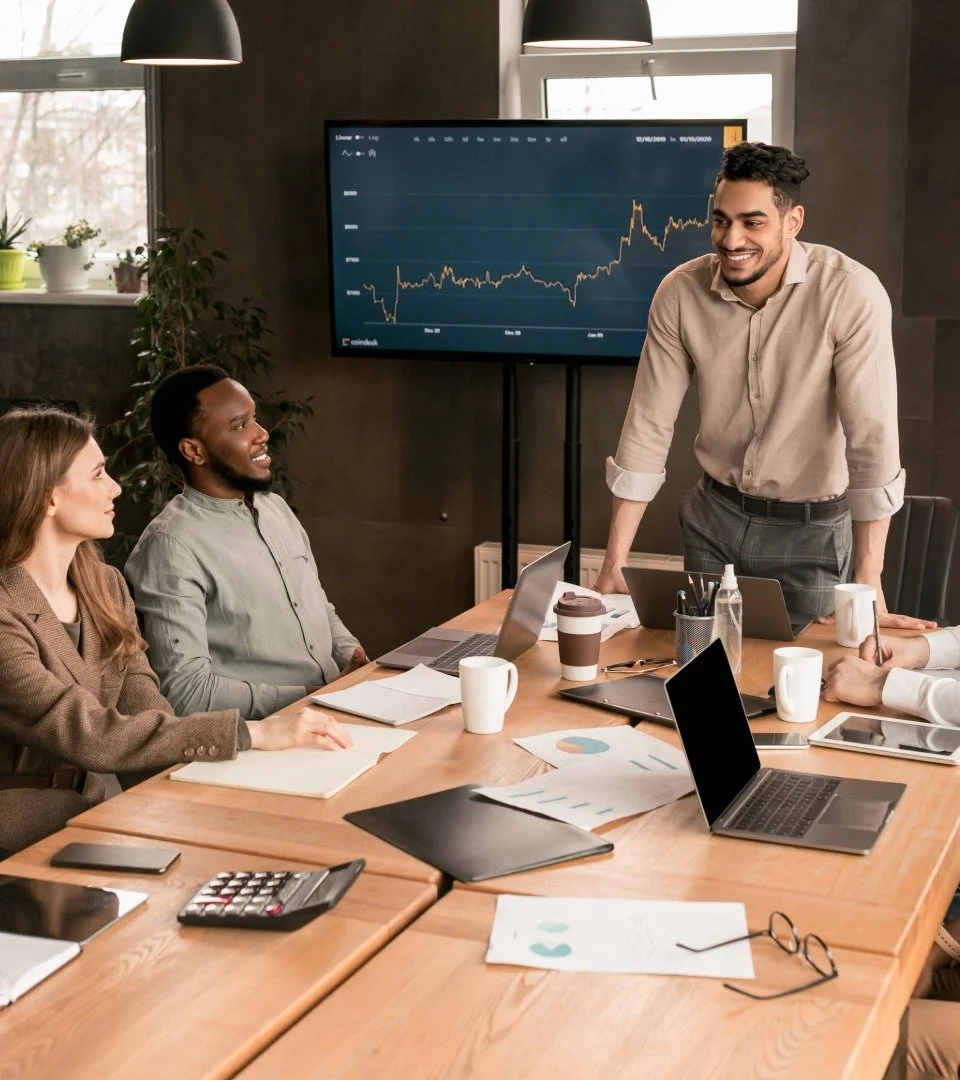 A diverse group of professionals in a meeting room with laptops, notebooks, and coffee cups, listening to a man presenting in front of a large screen displaying a financial chart.