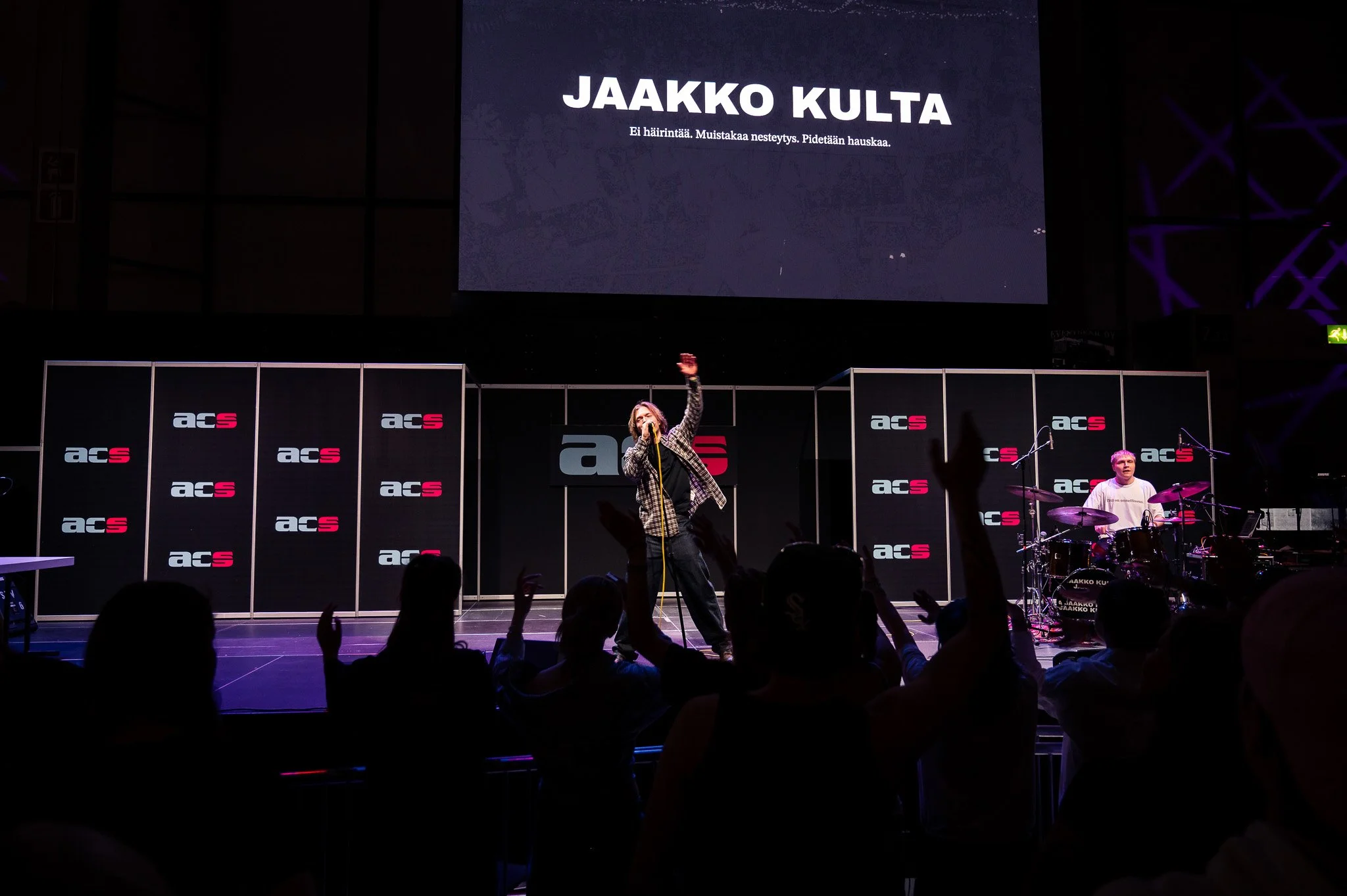 Esiintyjä lavalla, yleisöä etualalla, taustalla näyttöruutu, jossa lukee "JAAKKO KULTA", bändin kanssa.