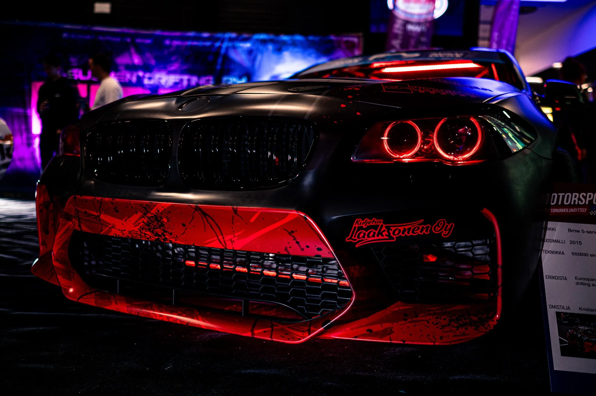 Musta urheiluauto esillä näyttelyssä, punaiset korostusvalot ja logo, taustalla ihmisiä ja värikkäitä valoja.