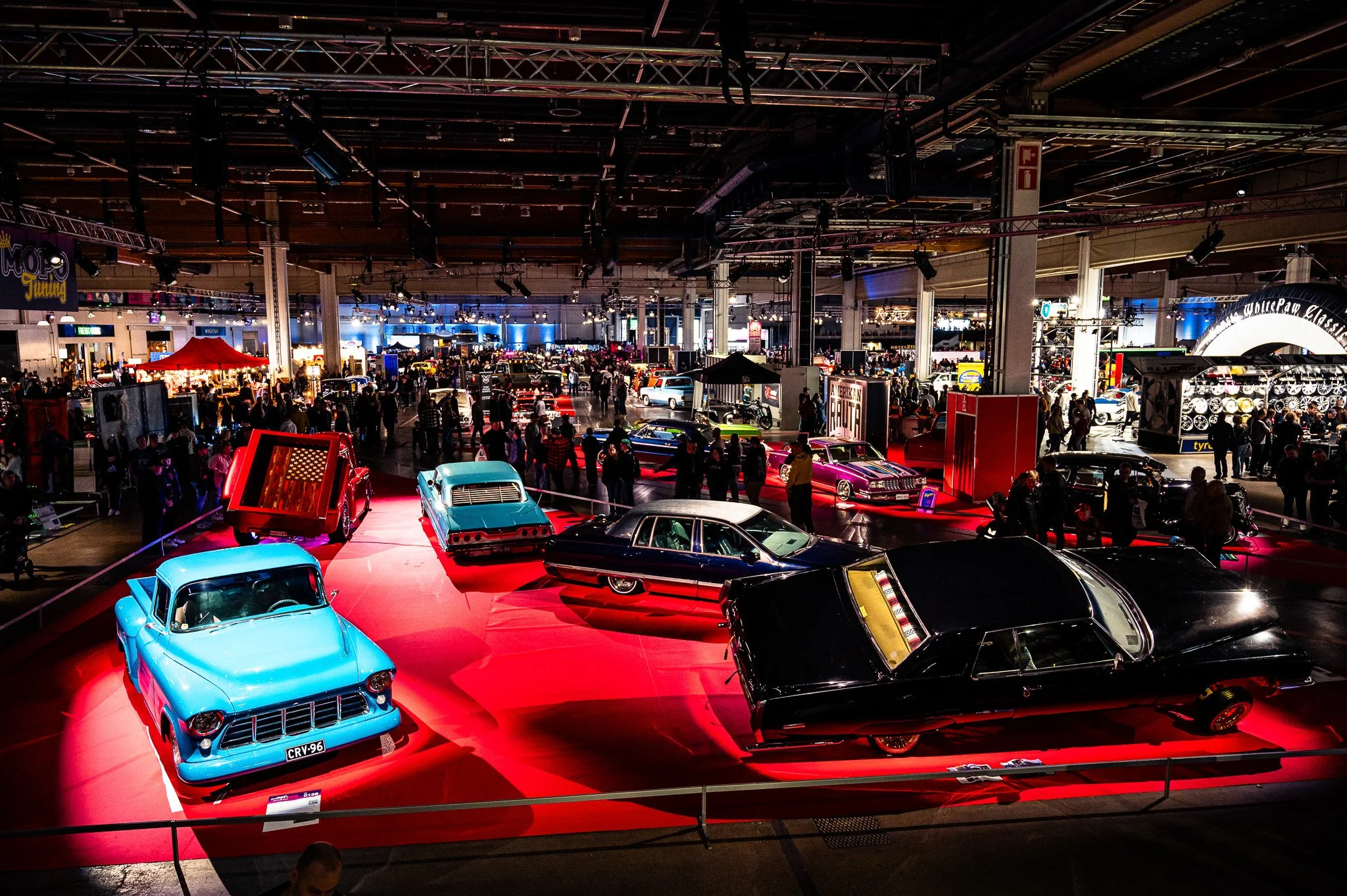 Näyttelyhallissa on esillä klassisia ja vintage-autoja, useita ihmisiä katselemassa autoja ja kulkuvälineitä, teemapisteitä ja näyttelytiskejä.