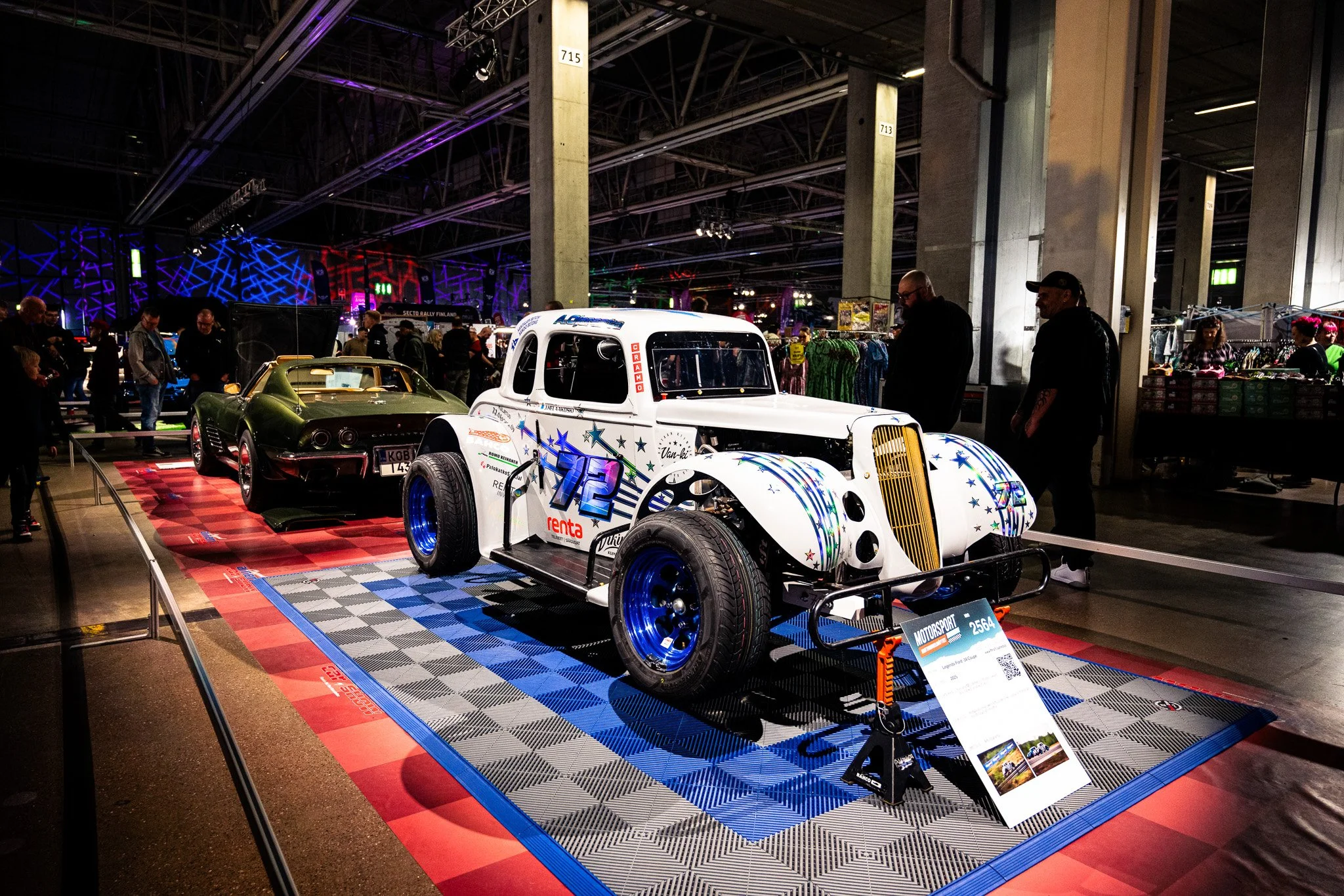 Näyttelytila, jossa kaksi erilaista autoa, yksi valkoinen ja toinen vihreä, esillä. Valkoinen auto on vanhanmallinen racing-auto, ja vihreä auto on urheiluauto. Taustalla on ihmisiä katsomassa autoja ja ostamassa tavaroita.