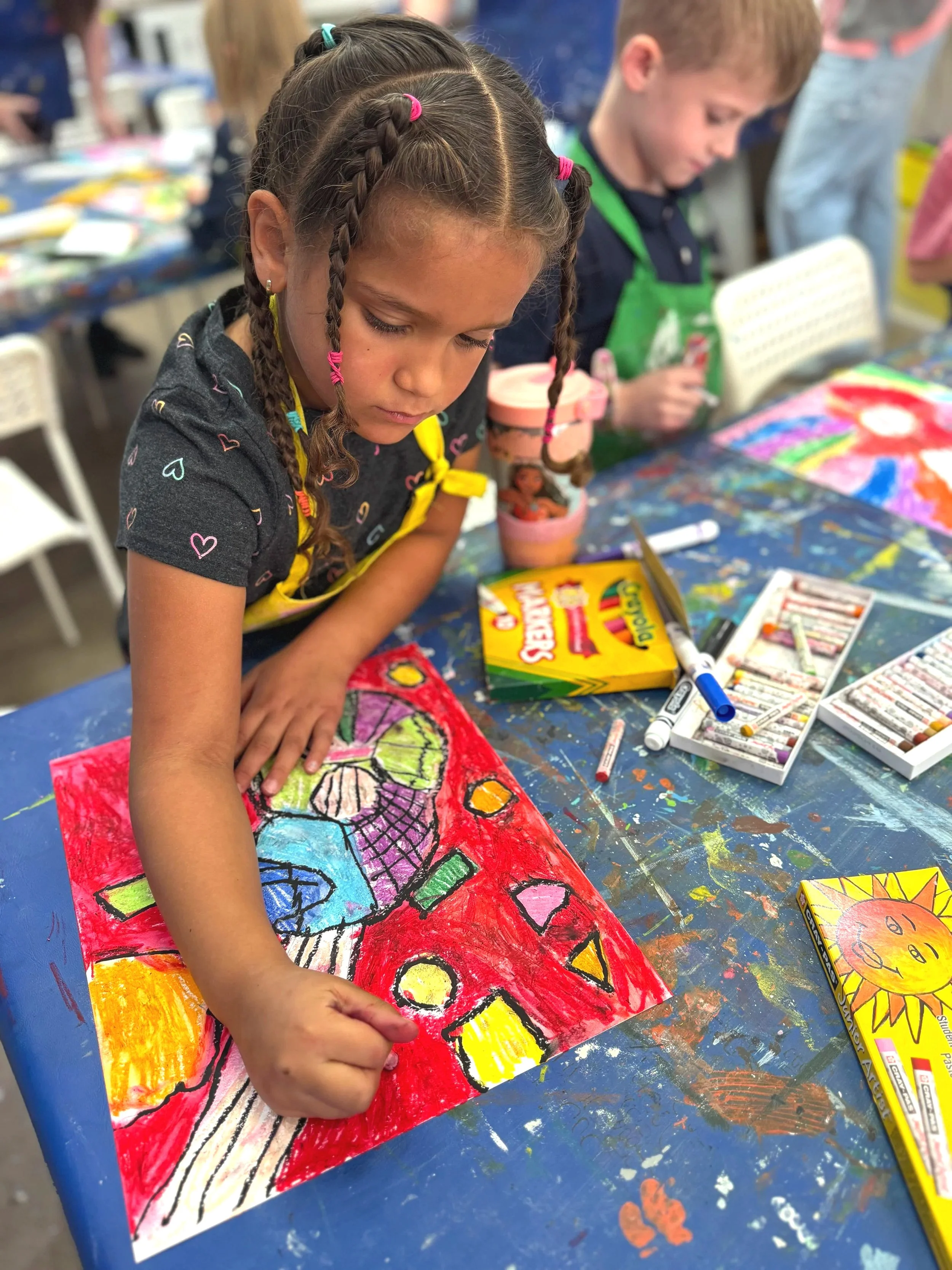 A young girl with braided hair and colorful hair ties is carefully coloring a vibrant abstract painting on paper with bright colors and bold patterns. In the background, a boy in a green apron is also engaged in the arts and crafts activity at a table covered with art supplies.