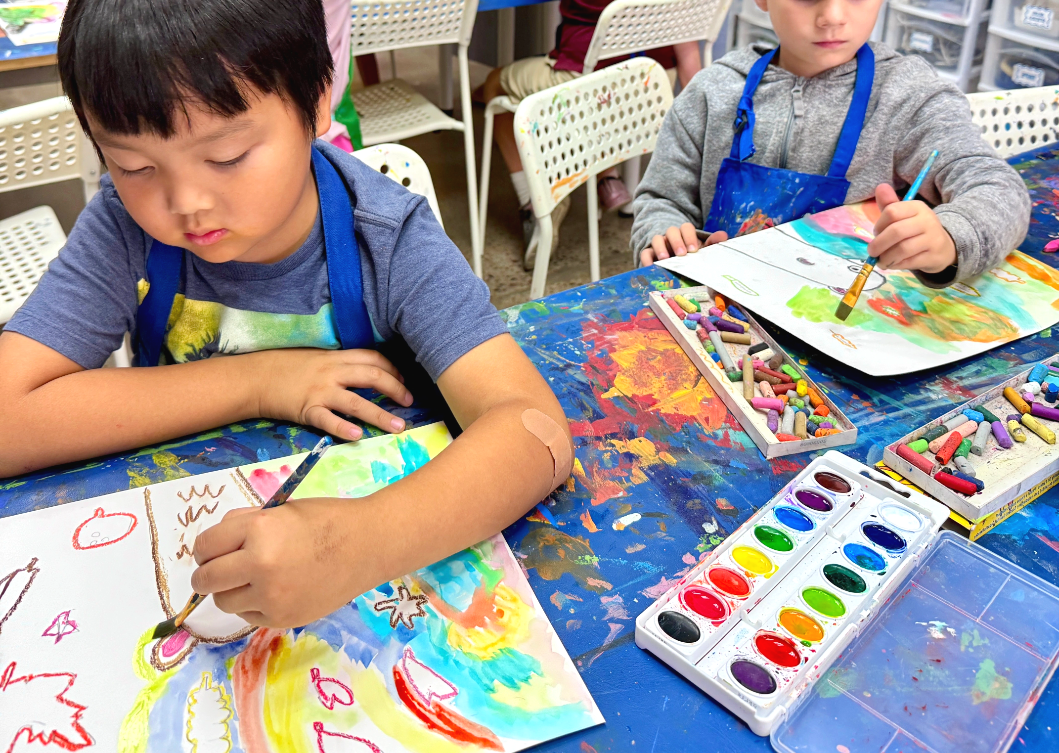 Two children painting with watercolors at a colorful art table. The children are focused on their artwork and wearing aprons.