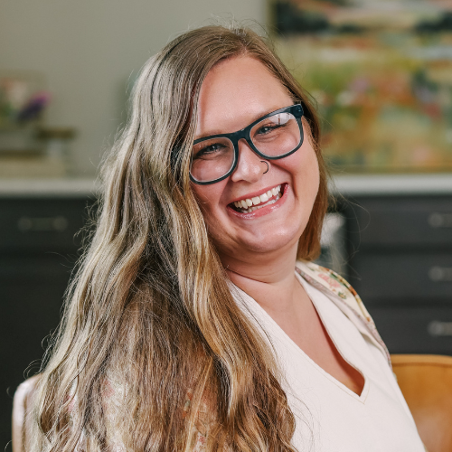 A woman smiling with long wavy hair and glasses in an indoor setting.