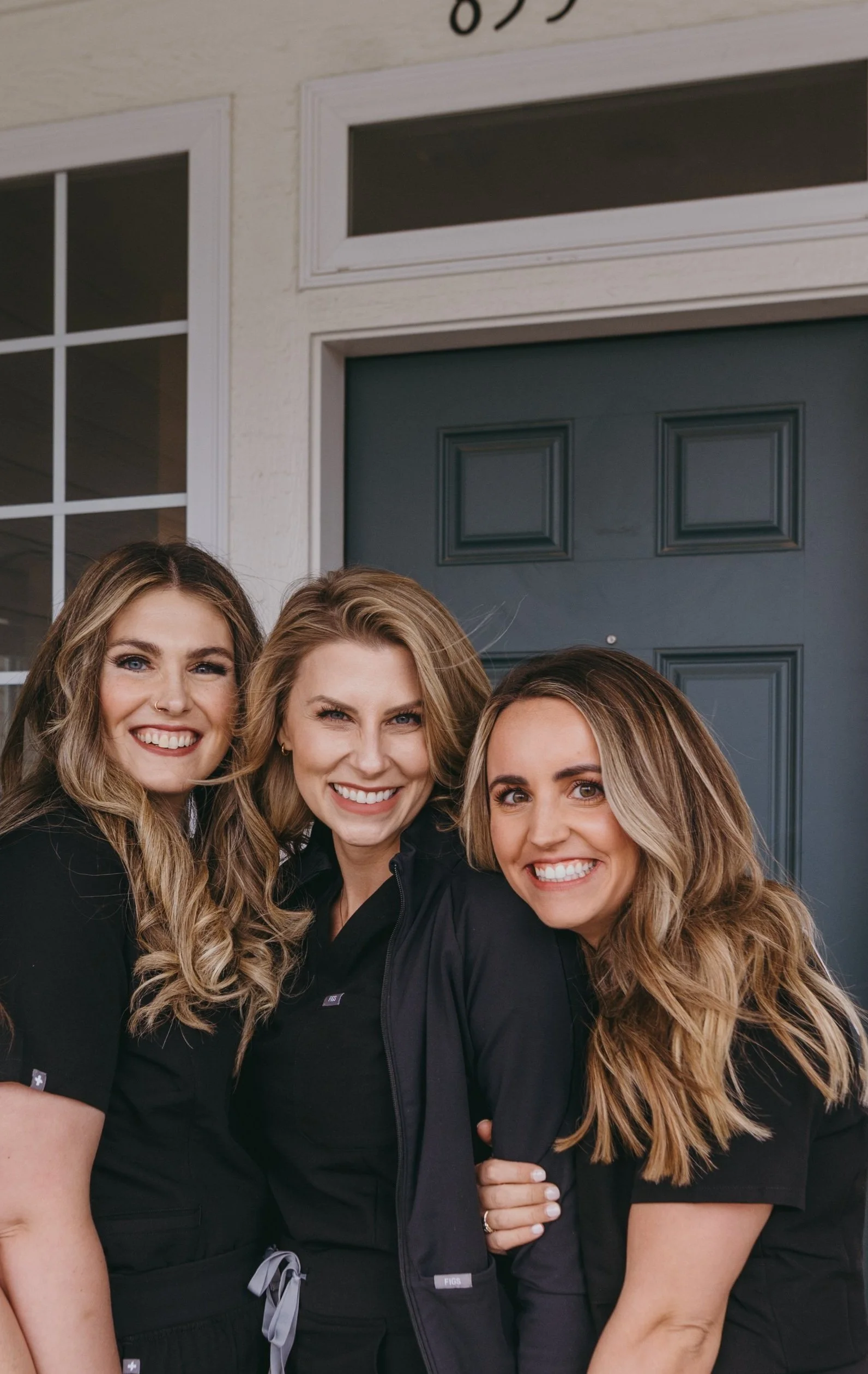 Three smiling women with long hair wearing black scrubs standing in front of a residential house with a blue door.