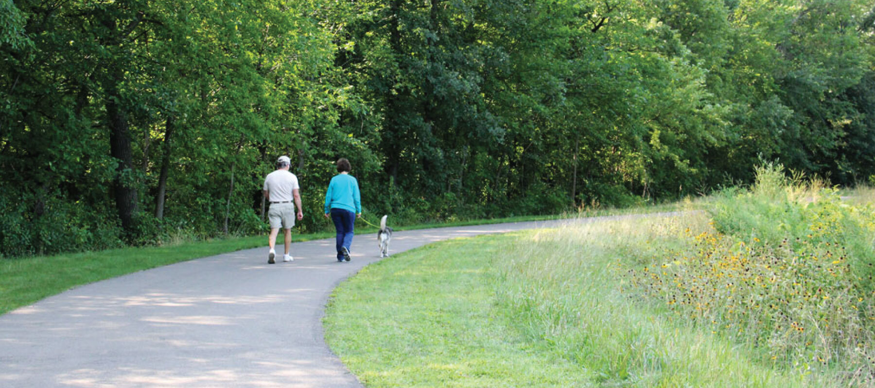 Photo of a man an woman walking a dog on a paved trail that runs through Coon Rapids Dam Regional Park. This trail is a part of the Mississippi River Regional Trail.