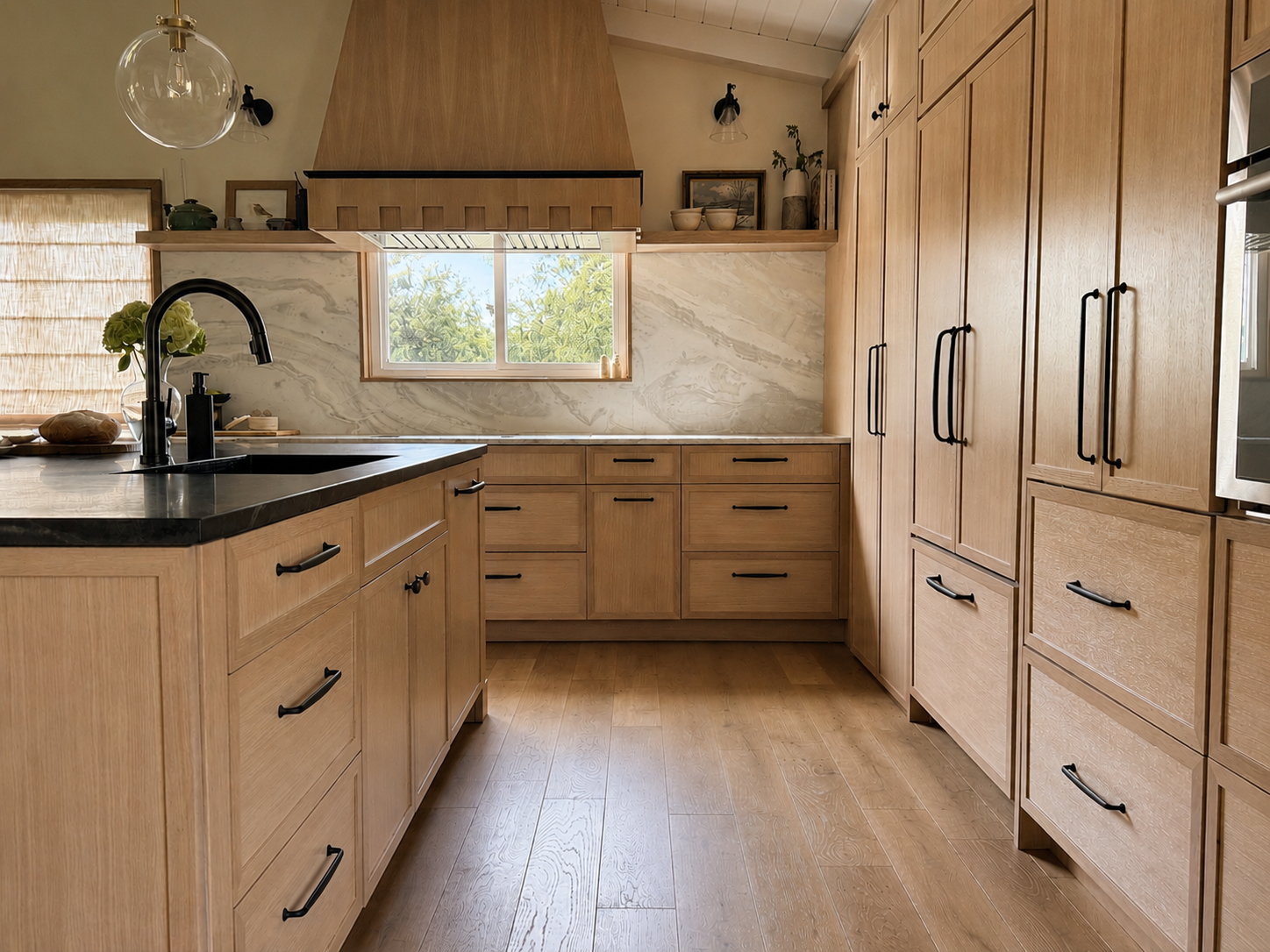 A kitchen with wooden cabinets, black handles, marble backsplash, black sink, faucet, a window with a view of trees, and a pendant light.