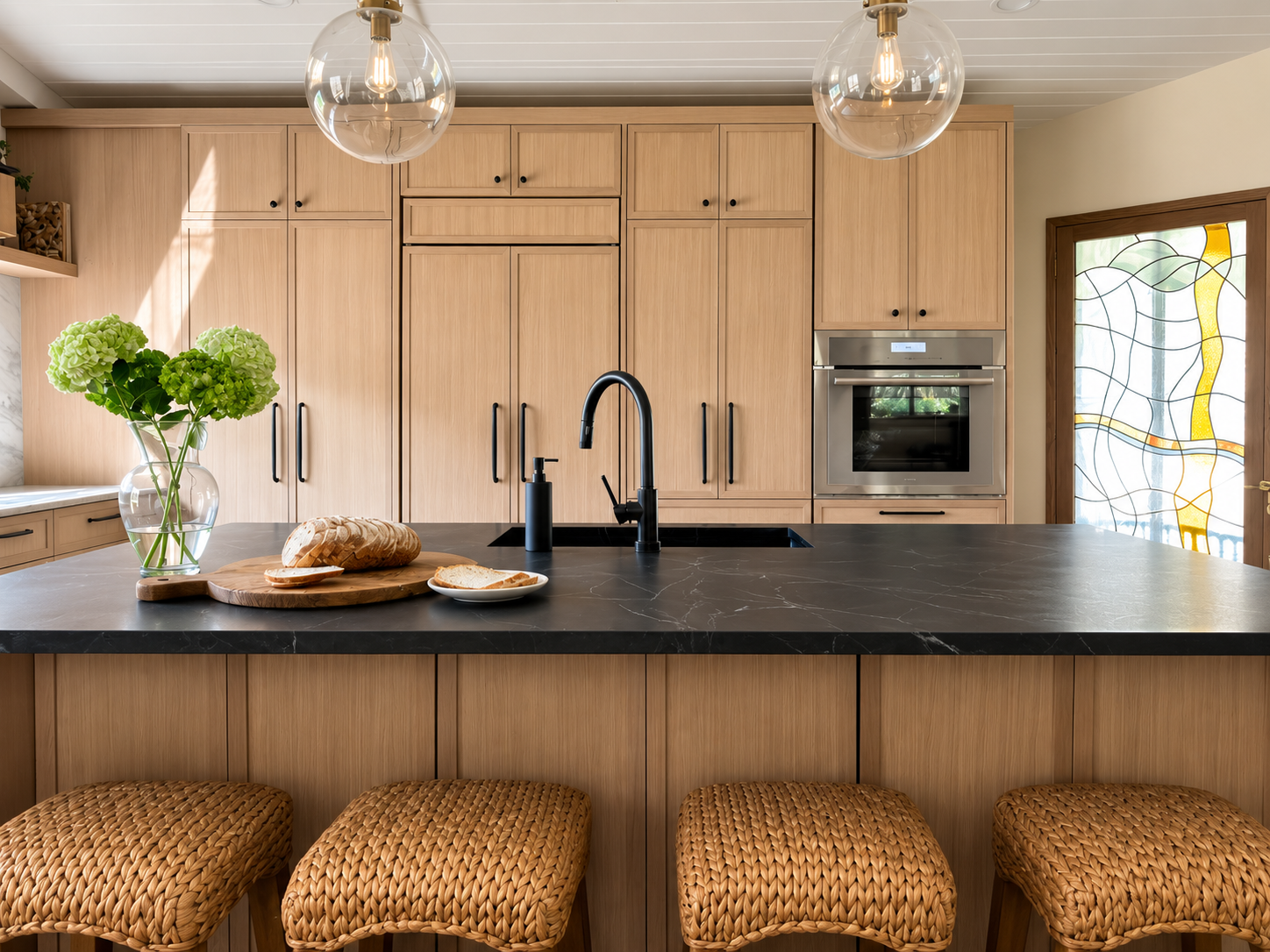 Modern kitchen with wooden cabinets, black countertop, and woven bar stools. A vase with green hydrangeas, bread, and slice of bread on a plate are on the counter. Two glass pendant lights hang from the ceiling. There is a built-in oven and a stained