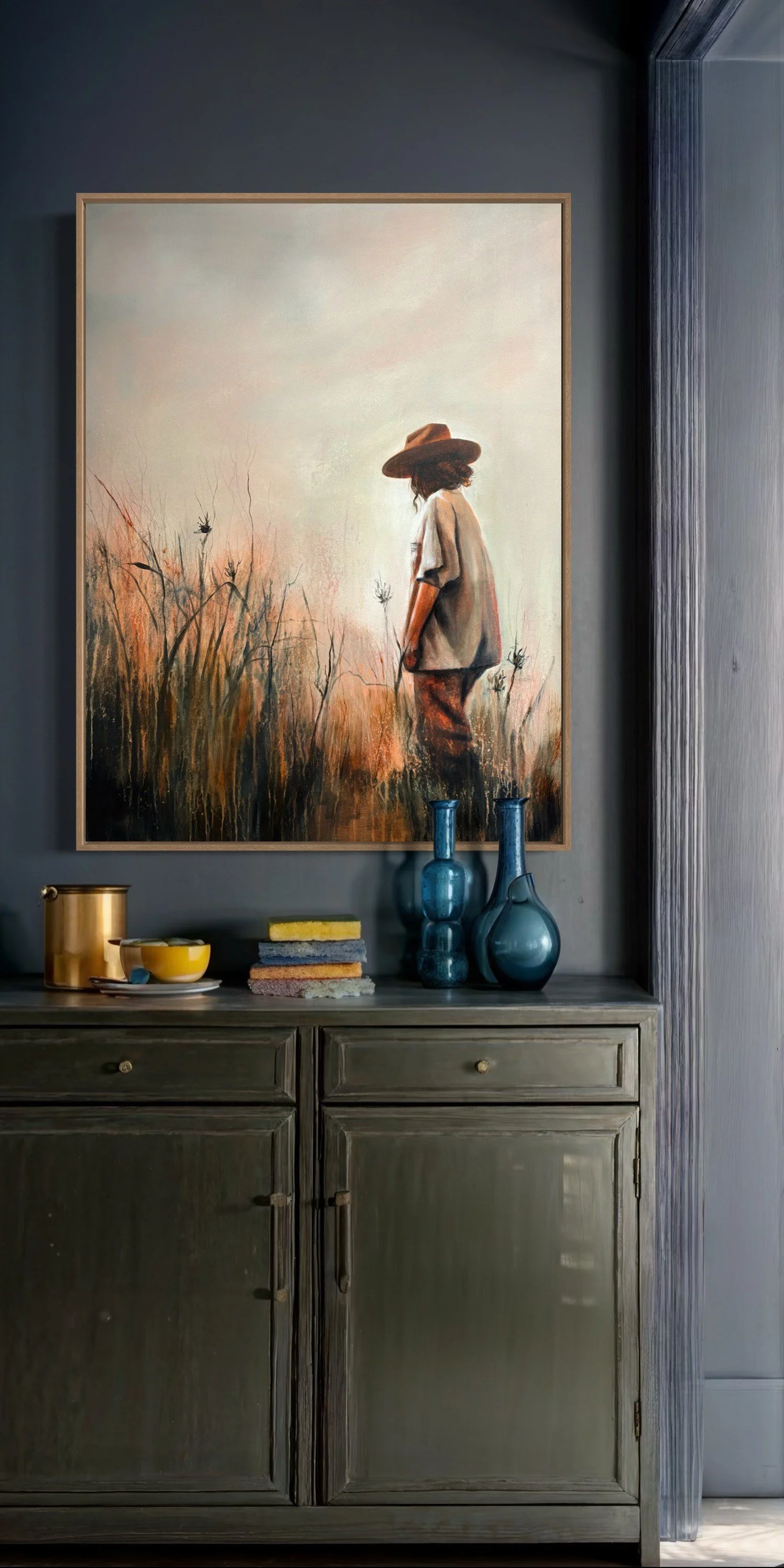 Painting of a woman in a hat standing in a field with dry grass and plants, behind a green cabinet with blue vases, a yellow bowl, and stacked sponges.
