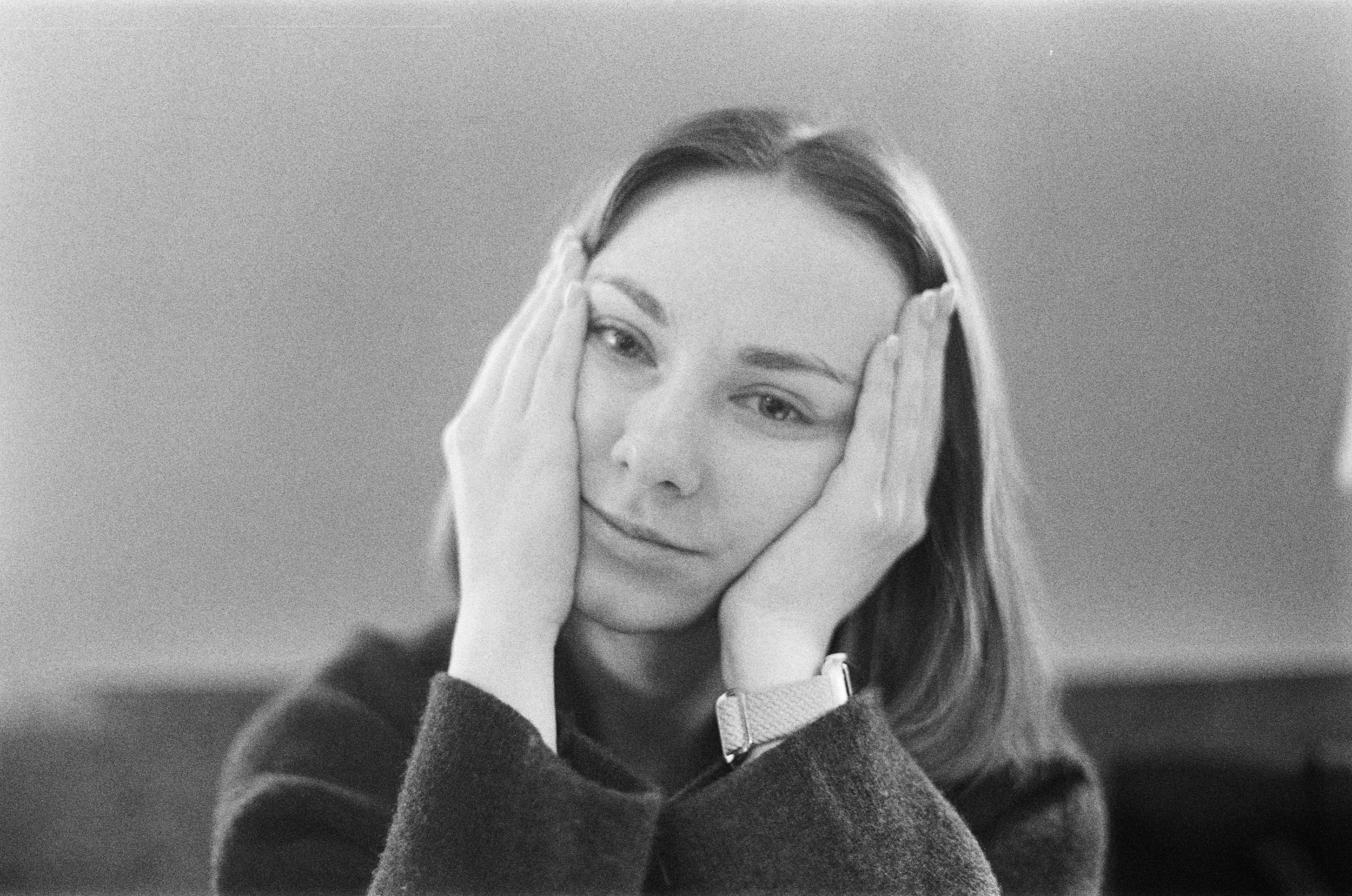 Une femme aux cheveux longs regarde pensivement, tenant son visage avec ses mains, arrière-plan neutre, en noir et blanc.