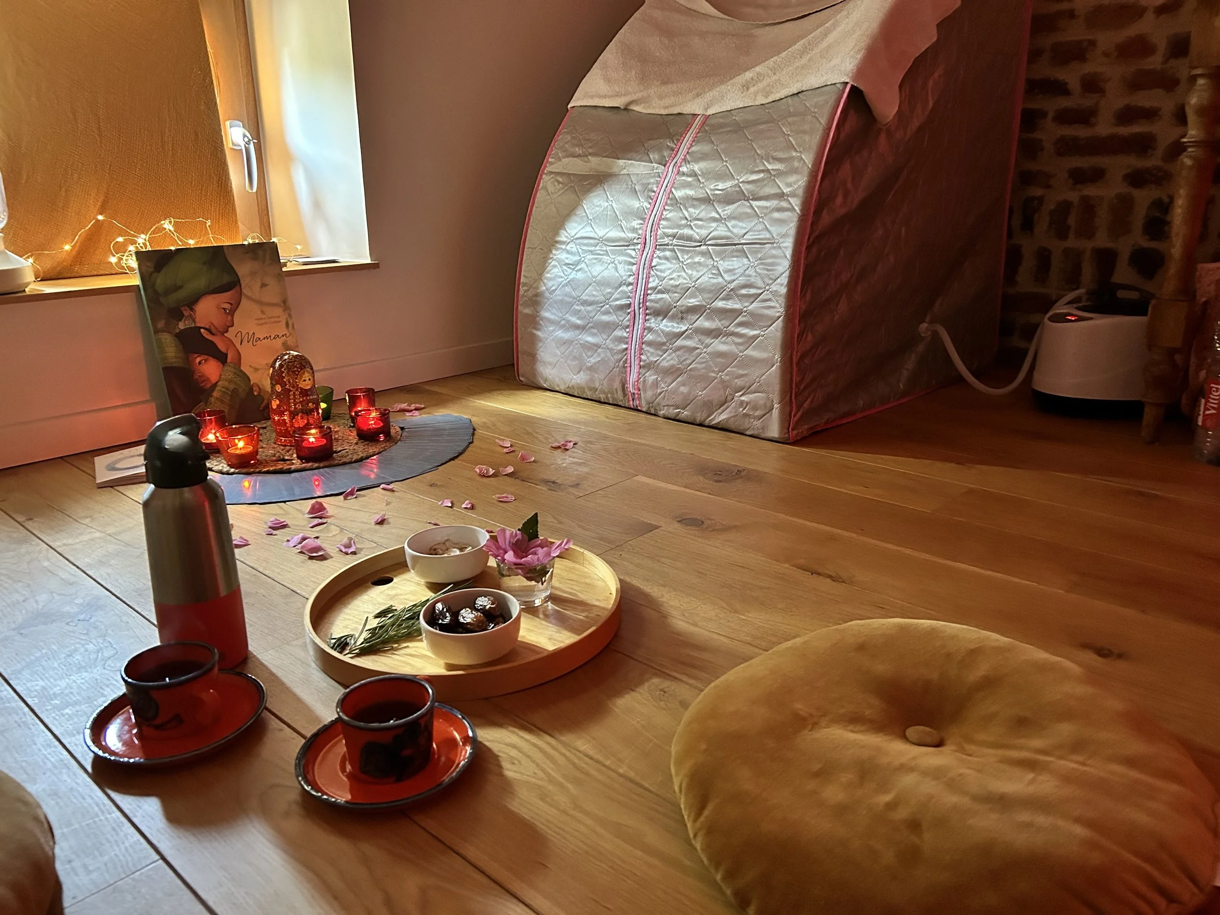 Un coin de chambre décoré pour une ambiance chaleureuse, avec des bougies rouges, des pétales de fleurs roses, un livre, une théière, des tasses et une nappe de midi, avec des lattes en bois pour le rituel rebozo.