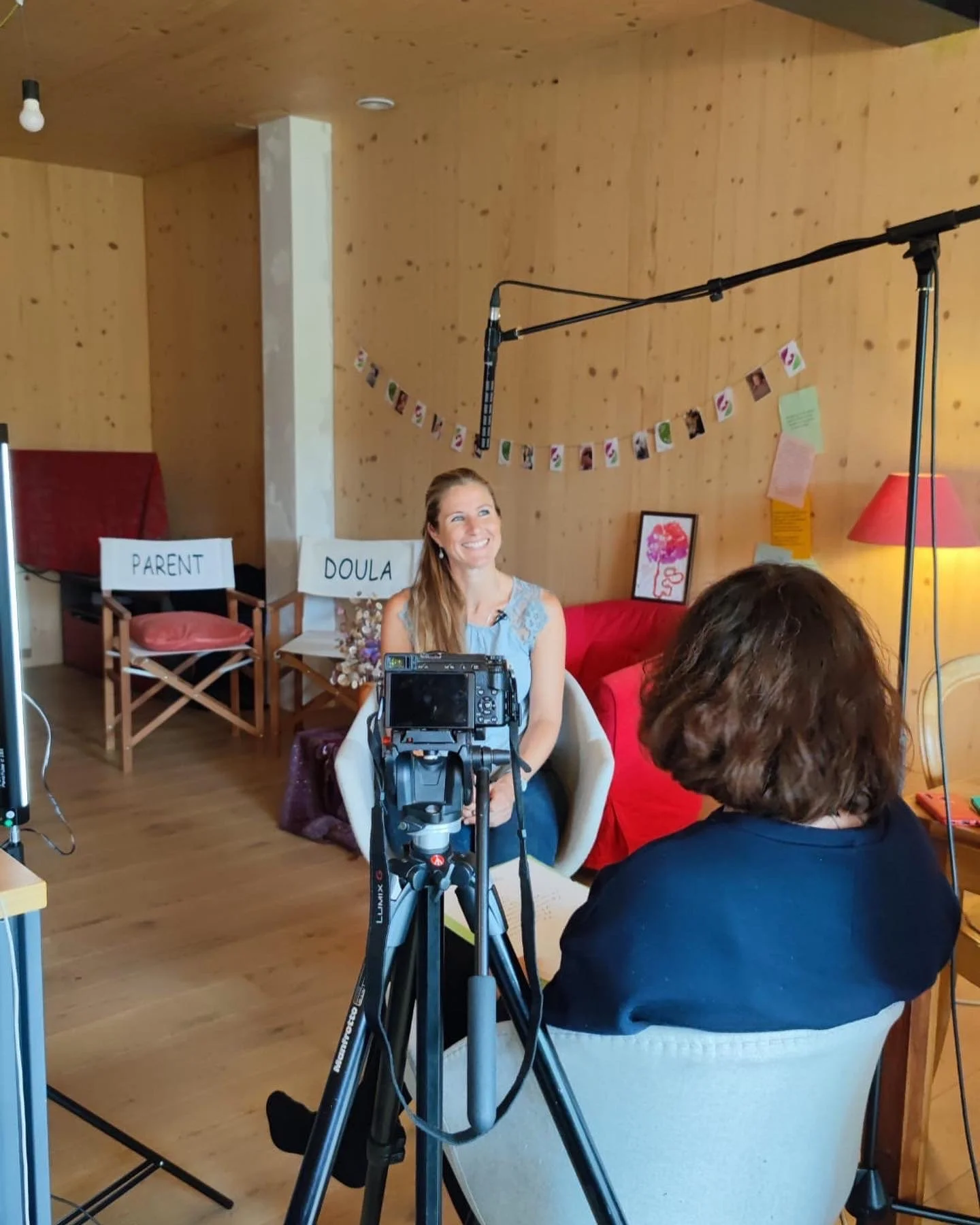 Une femme souriante étant interviewée dans une pièce en bois avec des décorations et des chaises, tandis qu'une personne avec des cheveux bruns assise devant elle filme avec une caméra.