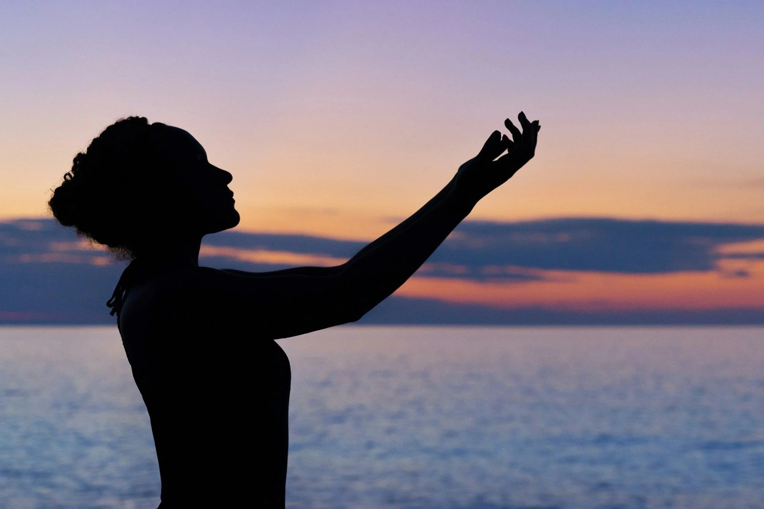 Silhouette d'une femme qui se tient près de l'eau, levant une main vers le coucher de soleil ou le lever du soleil.