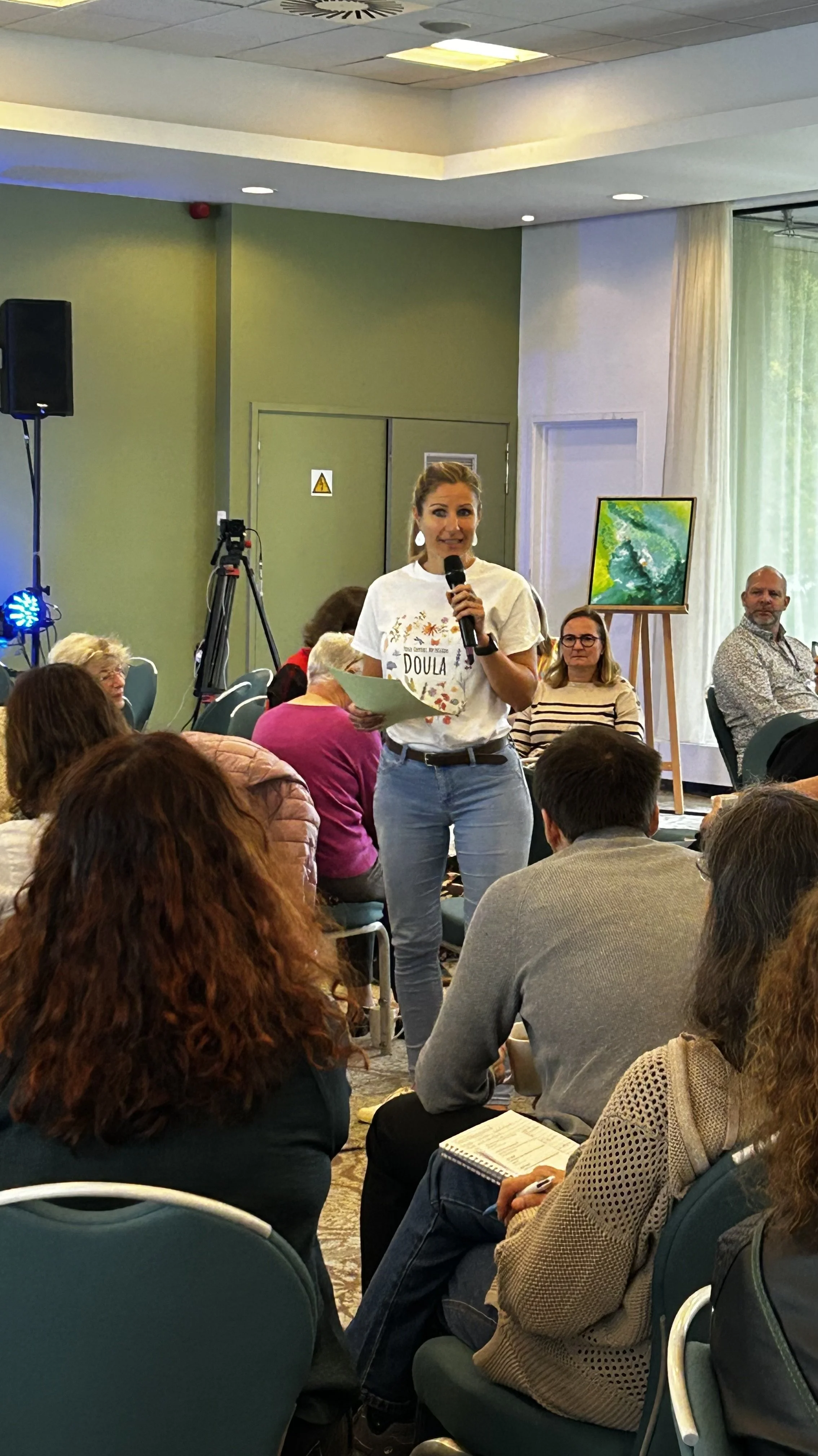 Une femme debout, tenant un microphone dans une main et un papier dans l'autre, s'adressant à un groupe de personnes assises lors d'une réunion ou d'une conférence. Le cadre est intérieur, avec une peinture sur un chevalet à l'arrière.