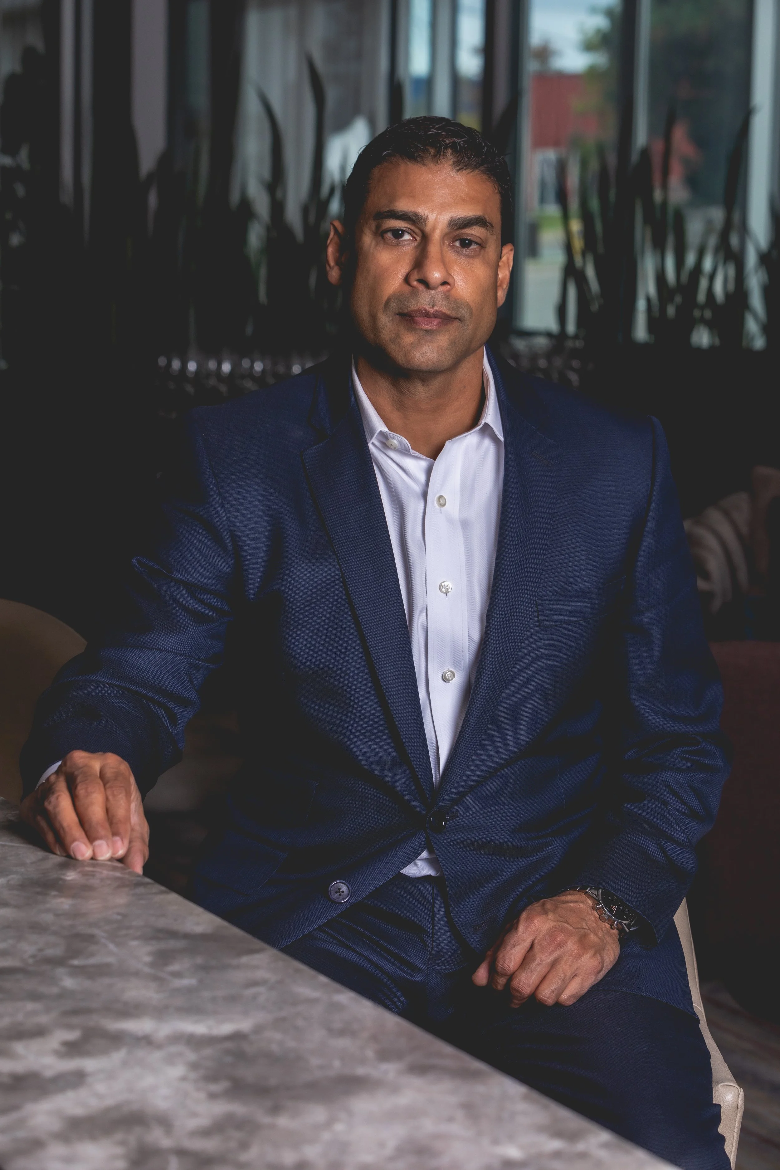 A man in a navy suit and white shirt sitting at a table in a modern space with large windows and plants in the background.