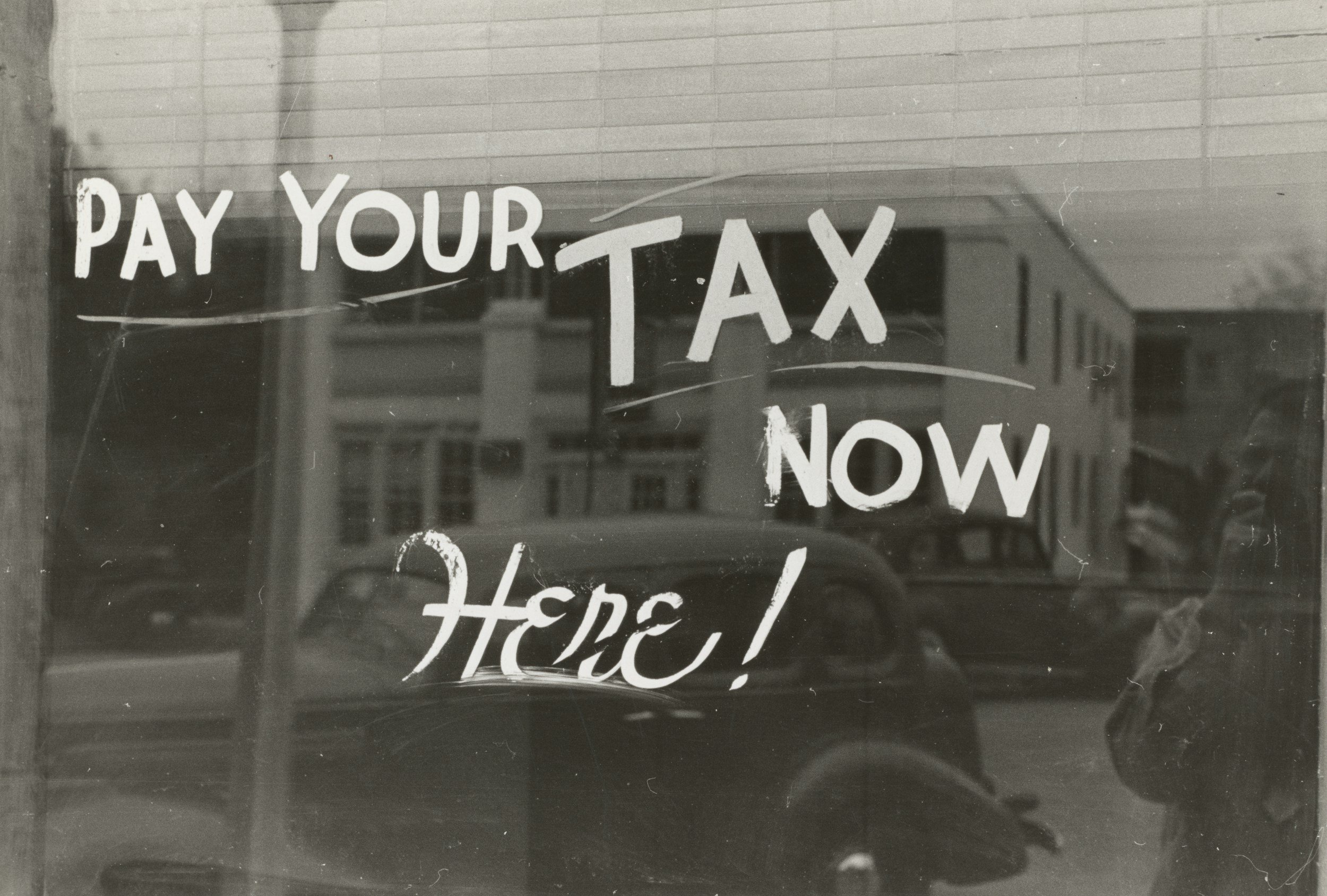 A sign on a glass door reads, 'PAY YOUR TAX NOW HERE!' with a reflection of buildings and cars outside in the background.