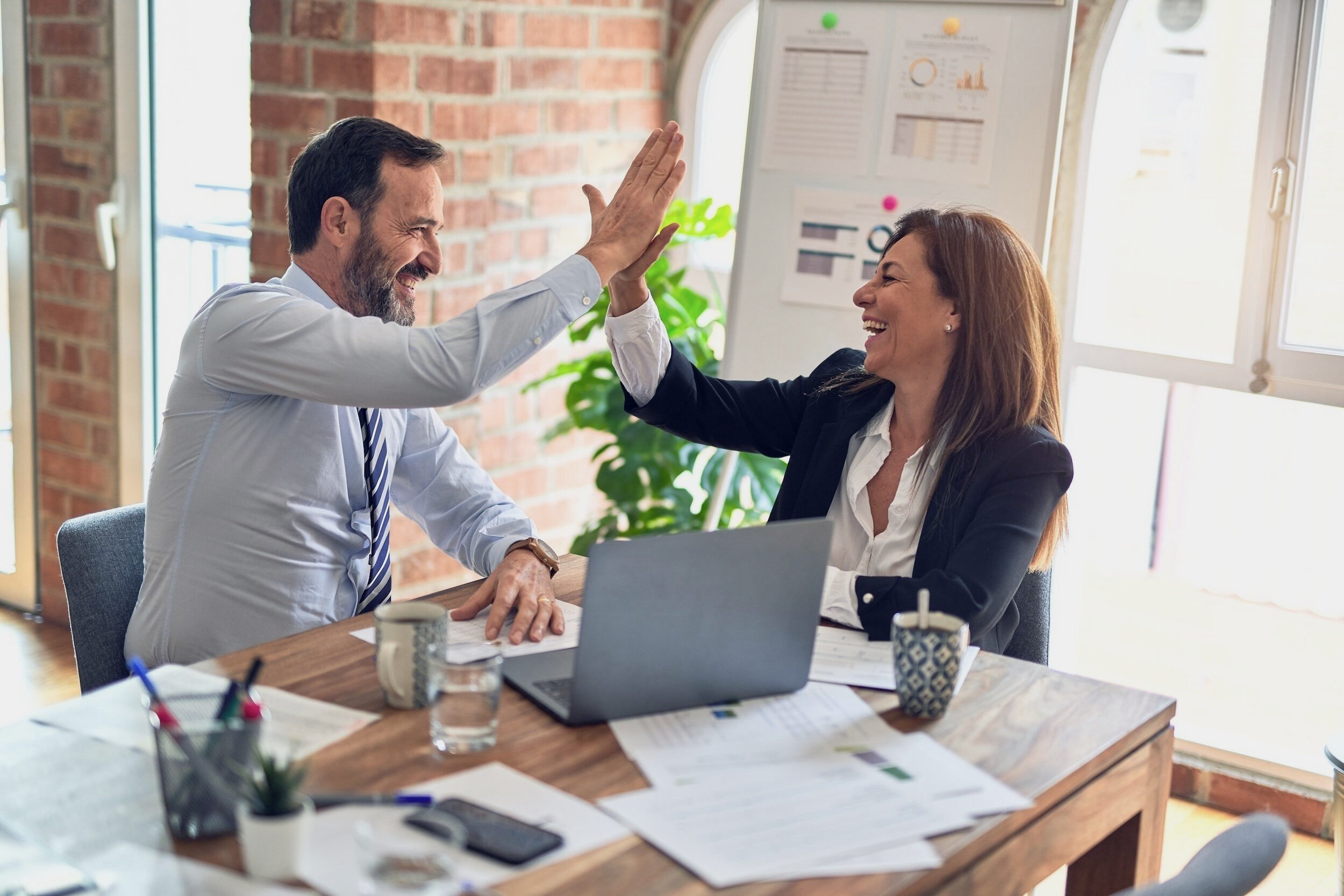 Two business professionals, a man and a woman, smiling and giving each other a high five in a bright office with a brick wall and large windows.