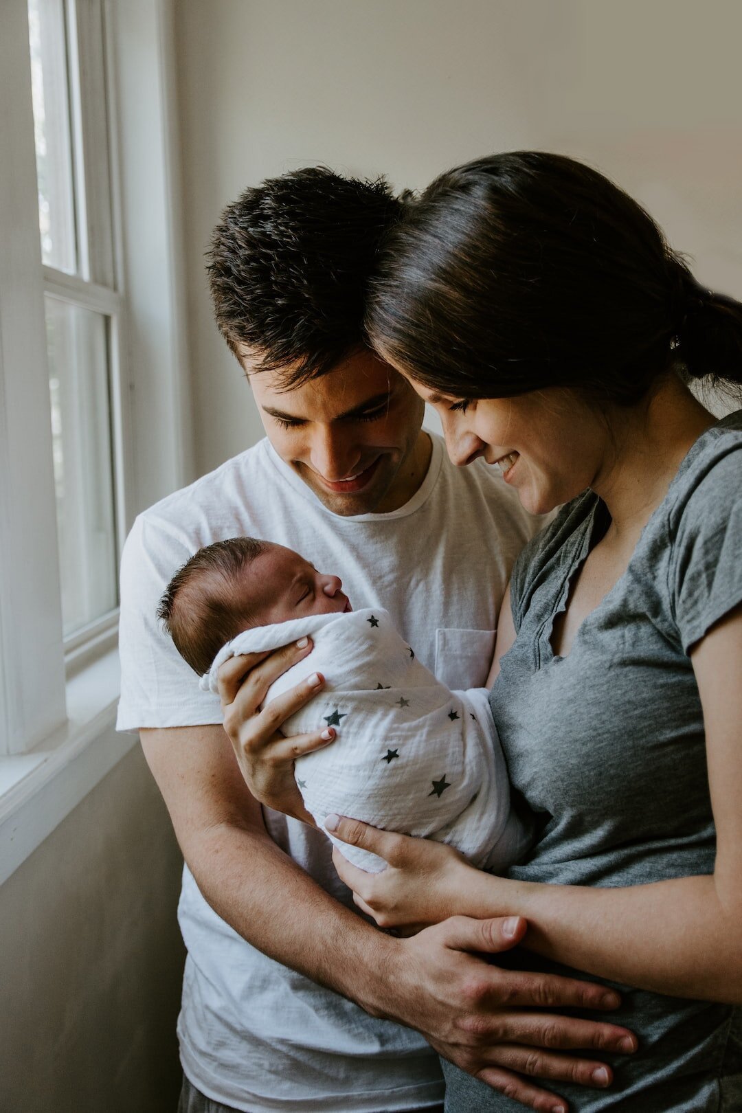 A happy young couple holding their newborn baby near a window.
