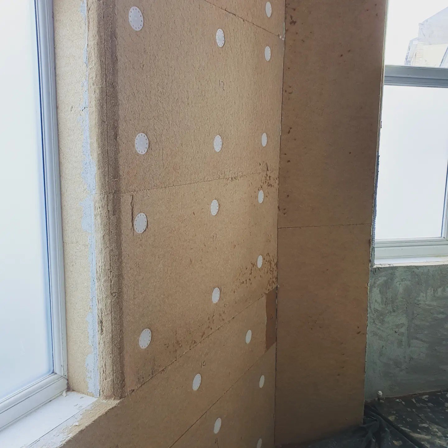 Interior wall under construction with drywall panels and round adhesive patches, next to a window.