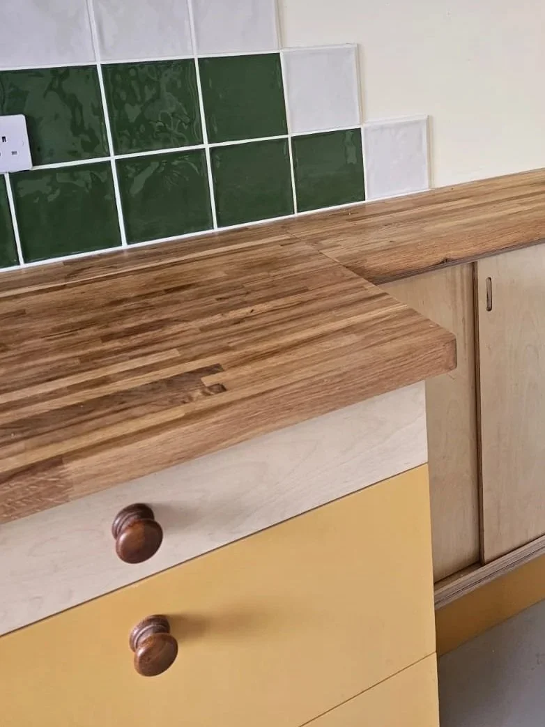 Close-up of kitchen cabinetry with a wooden countertop, a yellow drawer, and green and white tiled backsplash.