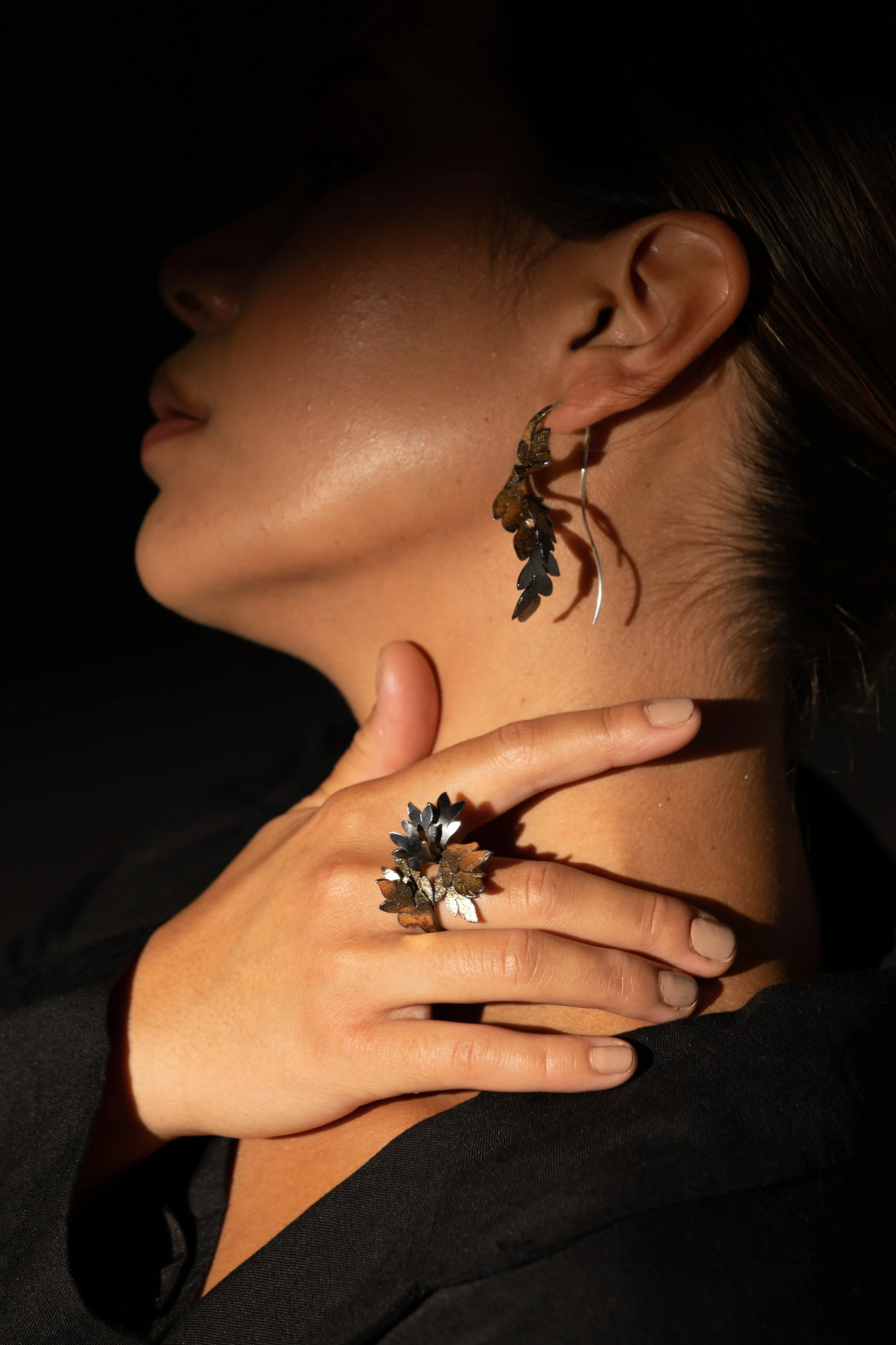 Close-up of a woman wearing black and gold leaf-shaped jewelry on her earlobe and ring, with her hand touching her neck, against a dark background.