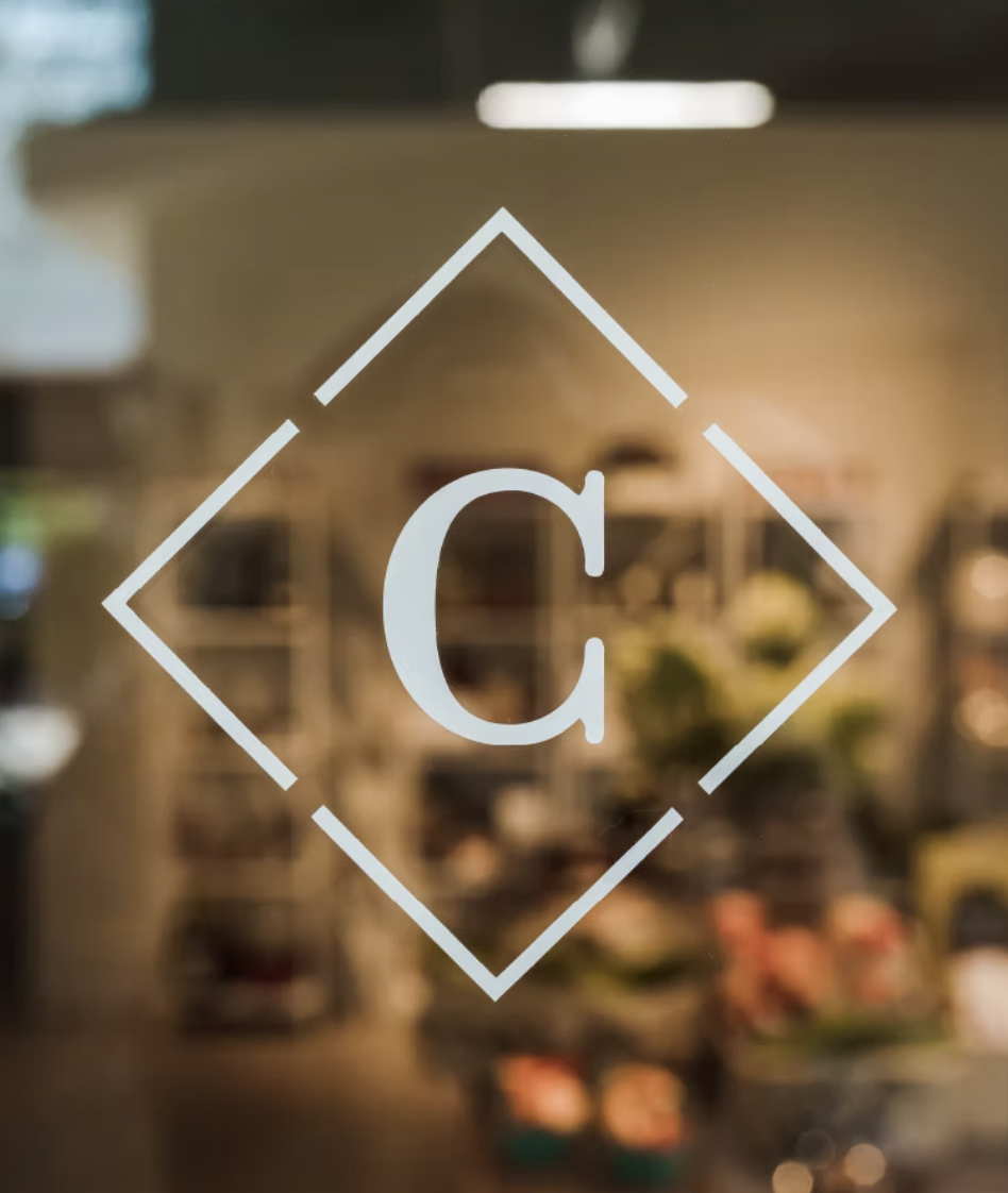 A close-up of a glass door with a logo featuring a large letter 'C' inside a diamond shape outline, with a blurred store interior in the background.