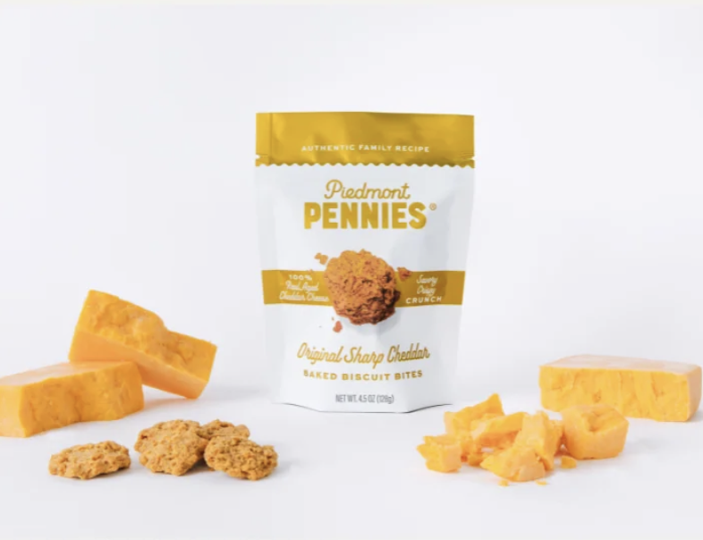 Bag of Piedmont Pennies baked biscuit bites with slices of cheddar cheese and cheddar cheese crumbles around it on a white background.