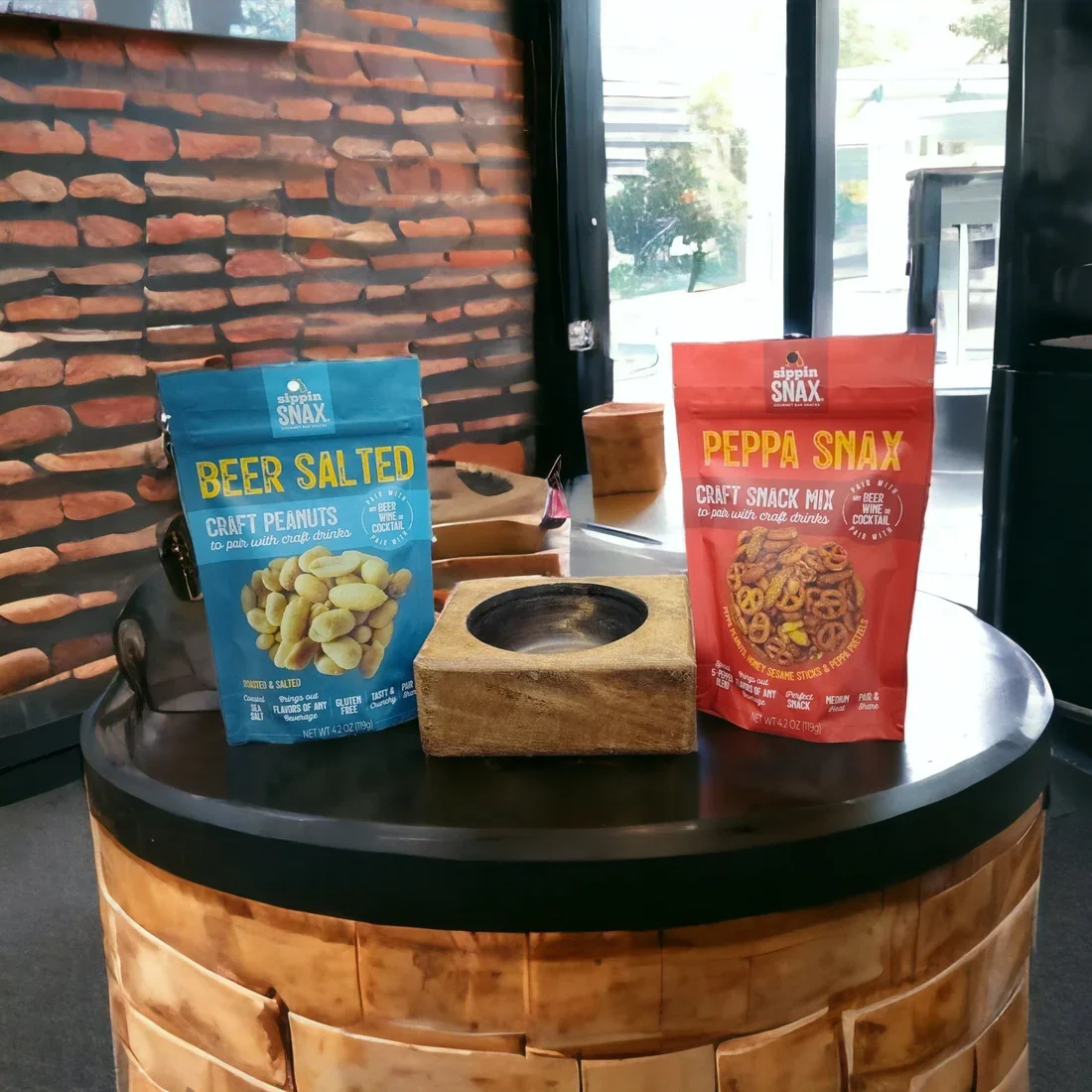 Two bags of craft snacks, one blue labeled 'Beer Salted Peanuts' and one red labeled 'Peppa Snax,' are placed on a round black table with a wooden base. A wooden tray or container is positioned behind the snacks, near a large window with natural light coming in, and a brick wall in the background.