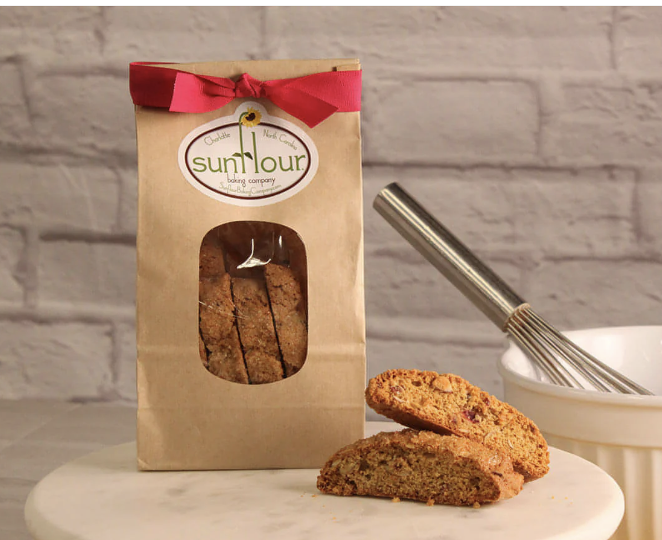 A brown paper bag of Sunflour baking company's cookies with a red ribbon on top, with two cookies placed in front of it on a white surface, and a cookie scoop resting on a white bowl in the background. The scene is set against a white brick wall.