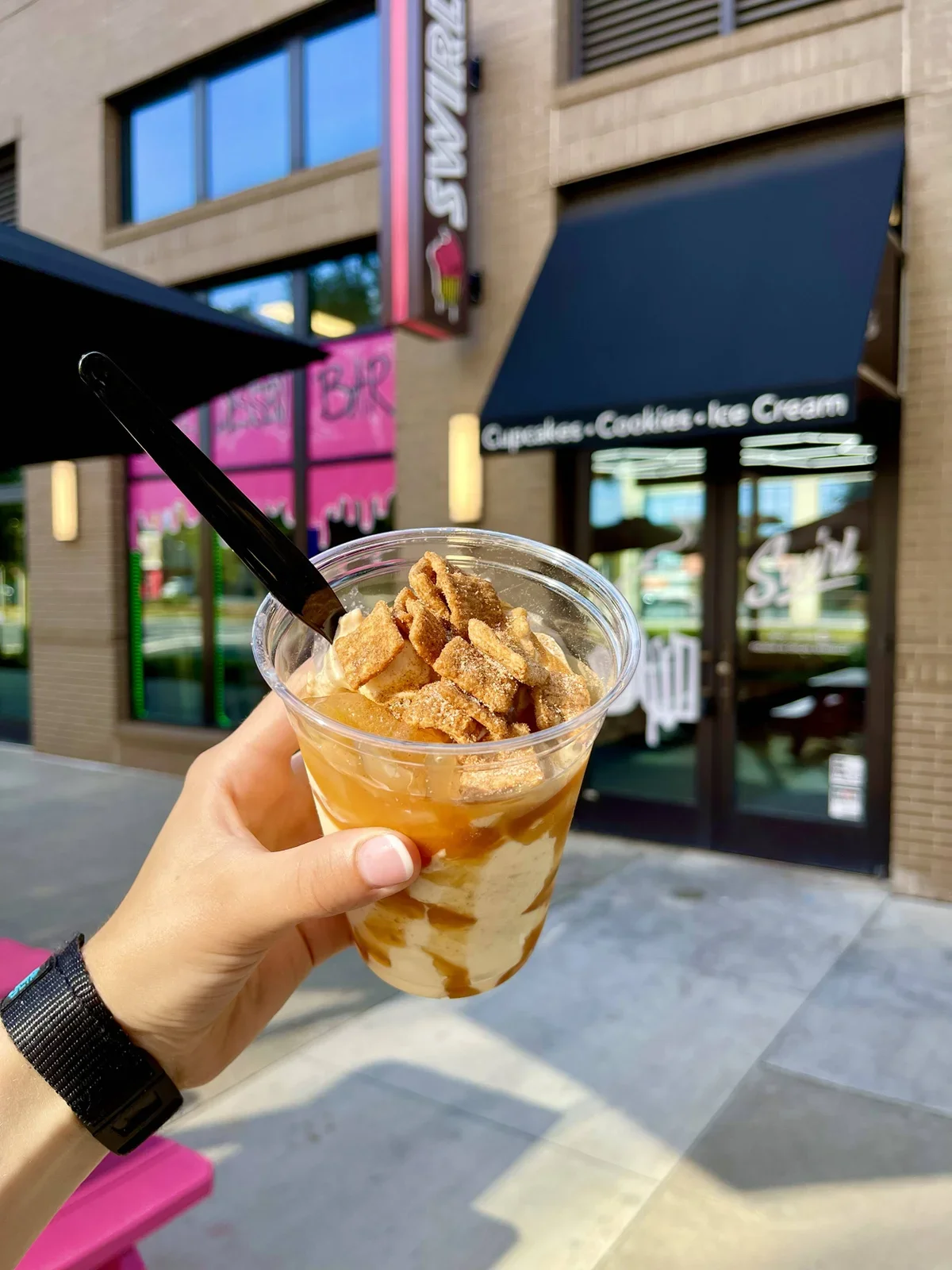 A hand holding a caramel-flavored iced coffee with whipped cream, caramel syrup, and pieces of caramel or toffee on top, outside a shop that sells cupcakes, cookies, and ice cream.