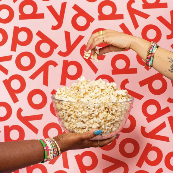 Two hands, one with a dark skin tone and the other with a light skin tone, holding a bowl of popcorn against a red and white background with the words 'pop' and 'yay' repeatedly printed, while the other hand is dropping popcorn into the bowl.