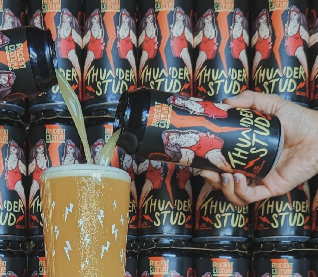 A hand holding a can of Thunder Stud energy drink with a hopped beer glass in the foreground, against a background of multiple cans with the same design, featuring a muscular woman in a red outfit and the text "Thunder Stud".