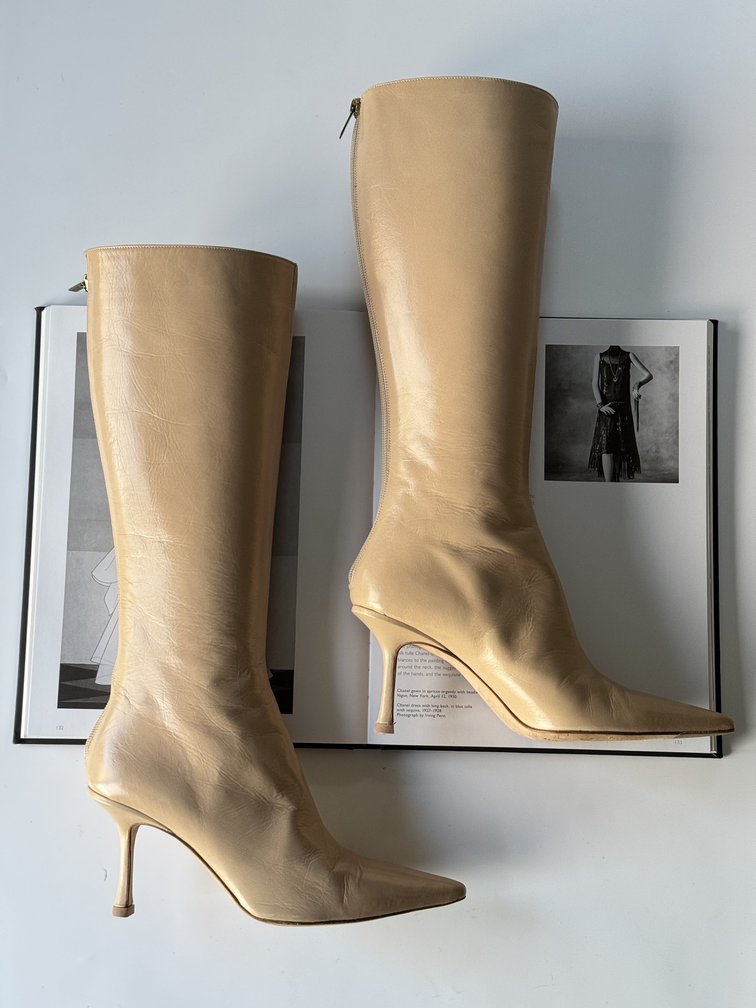A pair of beige high-heeled knee-high leather boots resting on an open magazine or book with black and white photographs.
