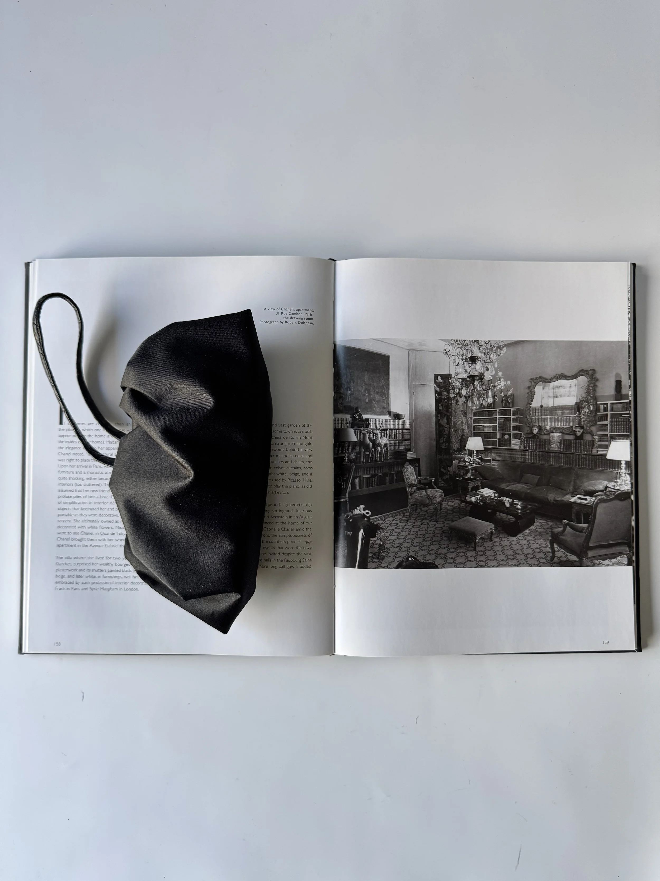 Open book with black face mask resting on the left page, black and white photograph of an ornate living room on the right page, plain white background.