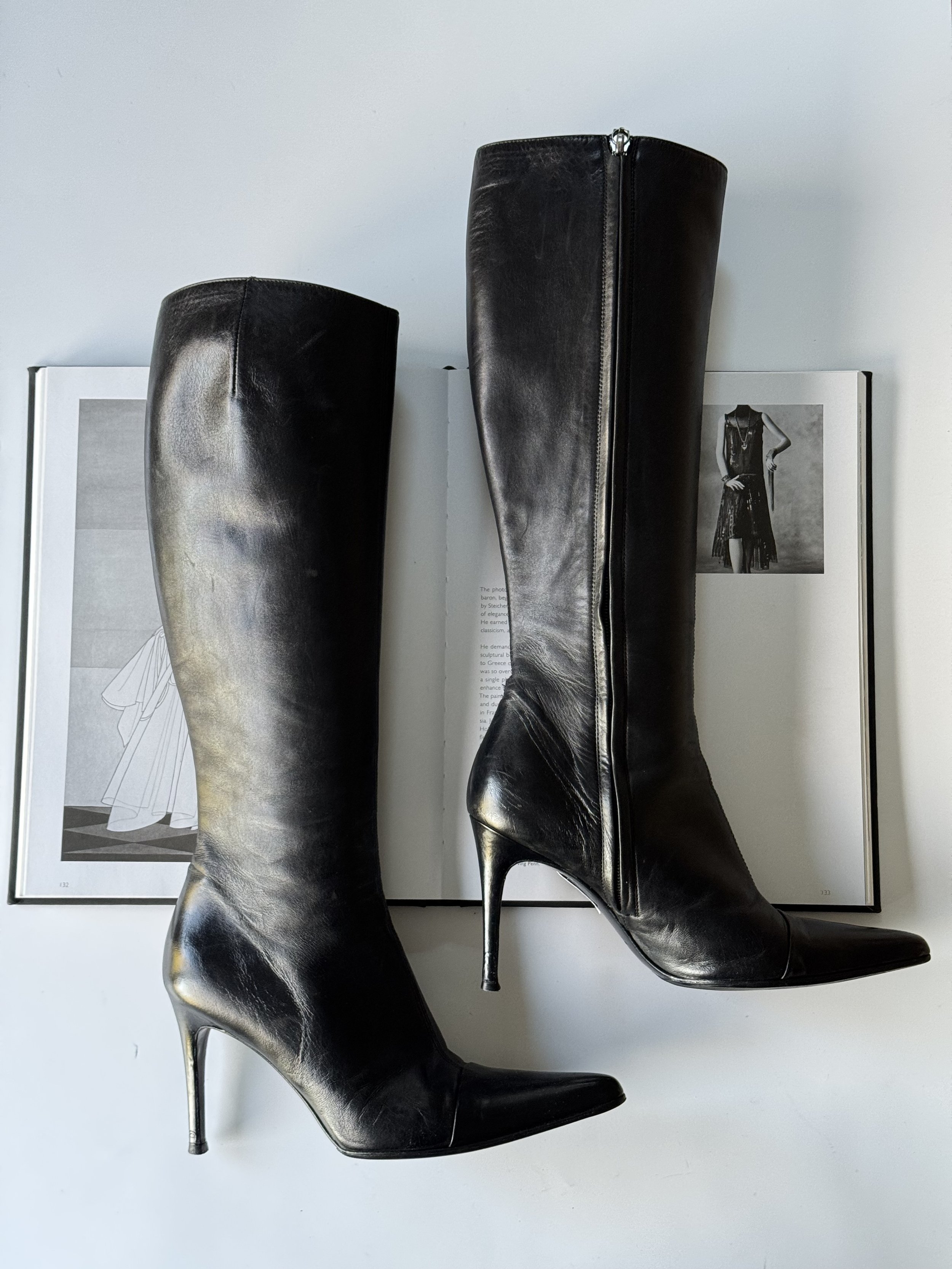 Pair of black high-heeled leather knee-high boots placed on an open magazine showing fashion images.