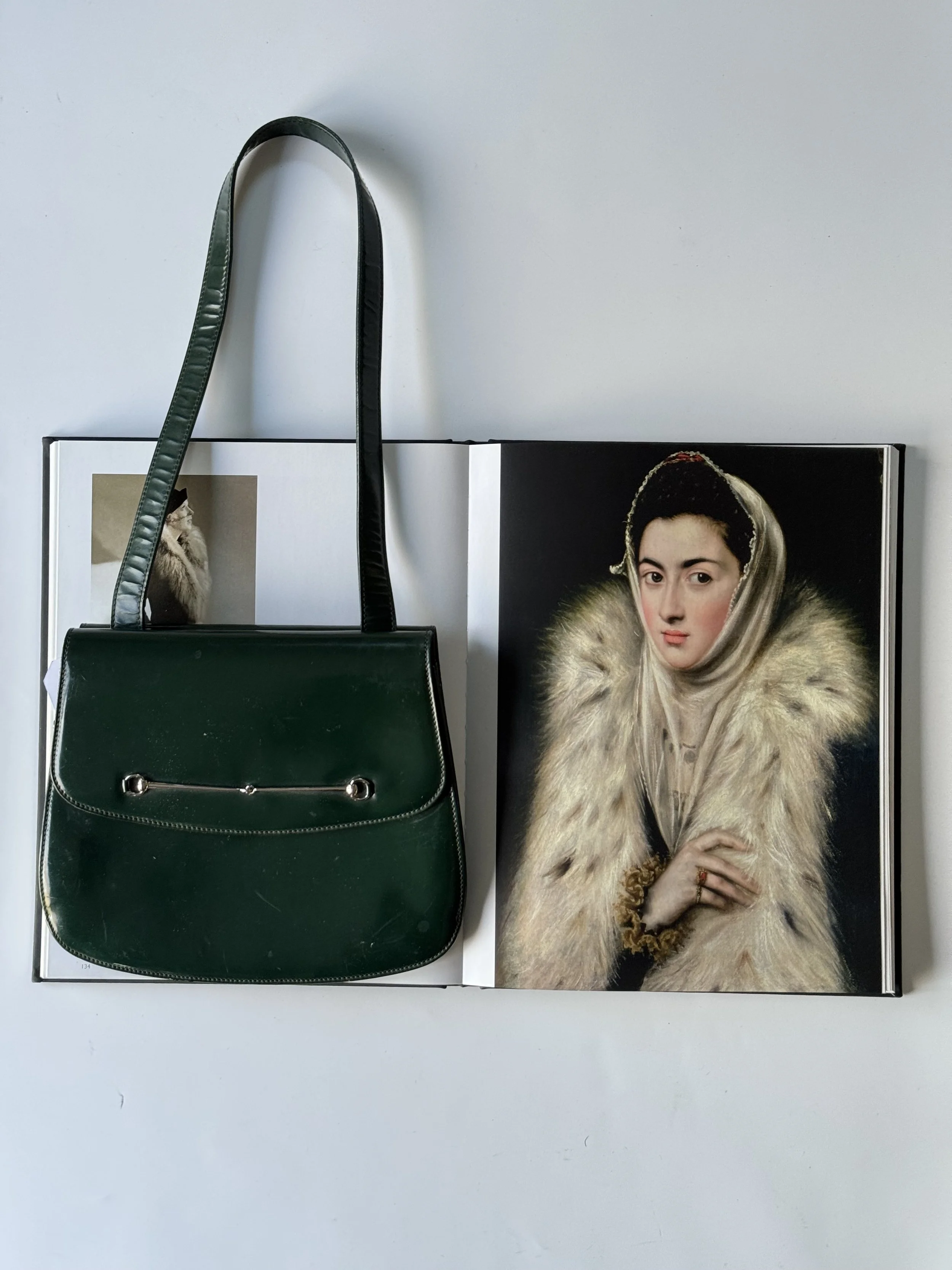 A green handbag hanging on the edge of an open book, which features a portrait of a woman with dark hair, wearing a fur coat and a headscarf.
