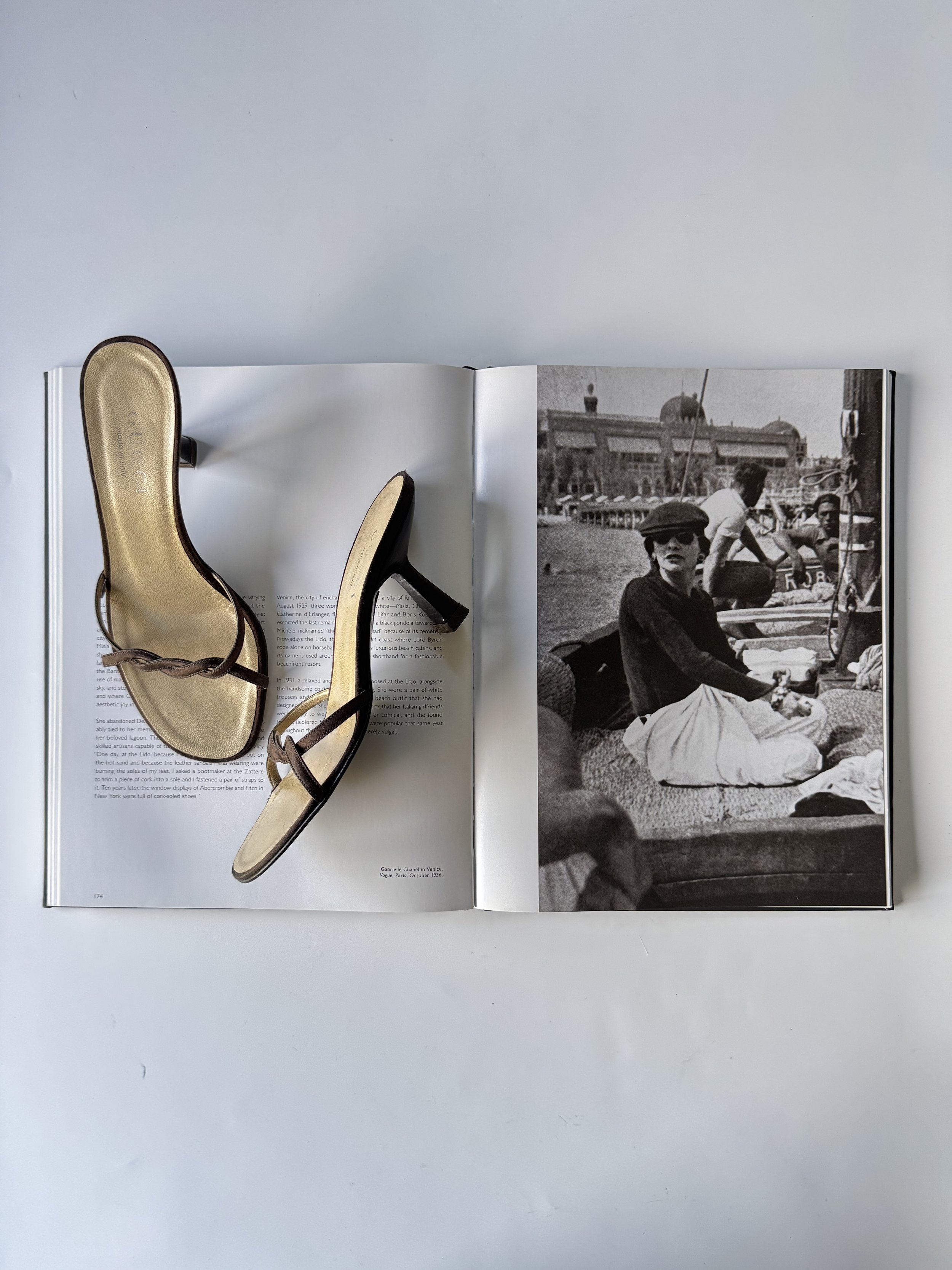 An open book with a pair of black strappy high-heeled sandals placed on the left page, and a black-and-white photograph of a woman sitting on a beach with other people and buildings in the background on the right page.