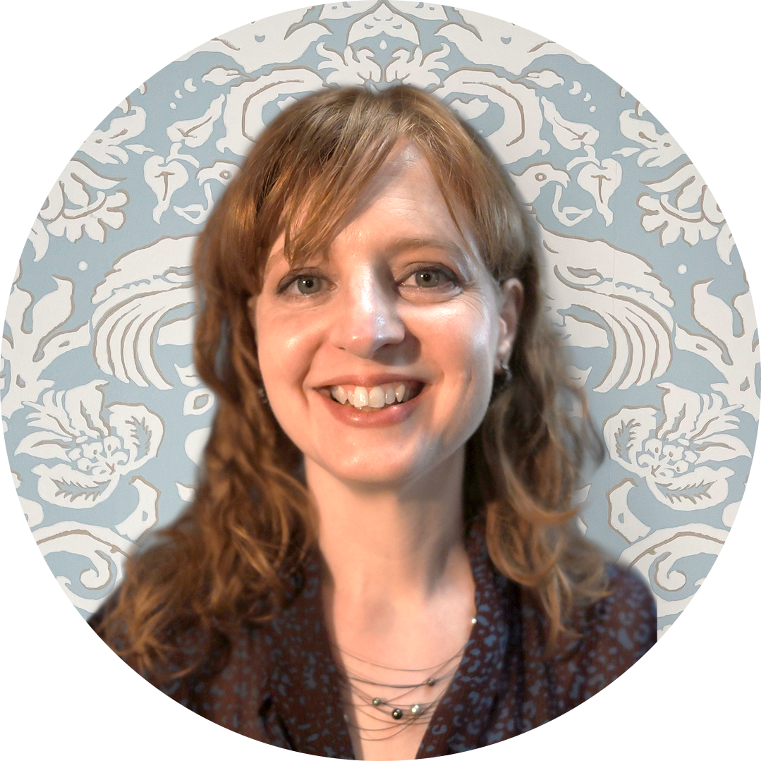 A woman with red hair and glasses smiling, wearing a shirt with a cat pattern, standing in front of a decorative blue and white wallpaper.