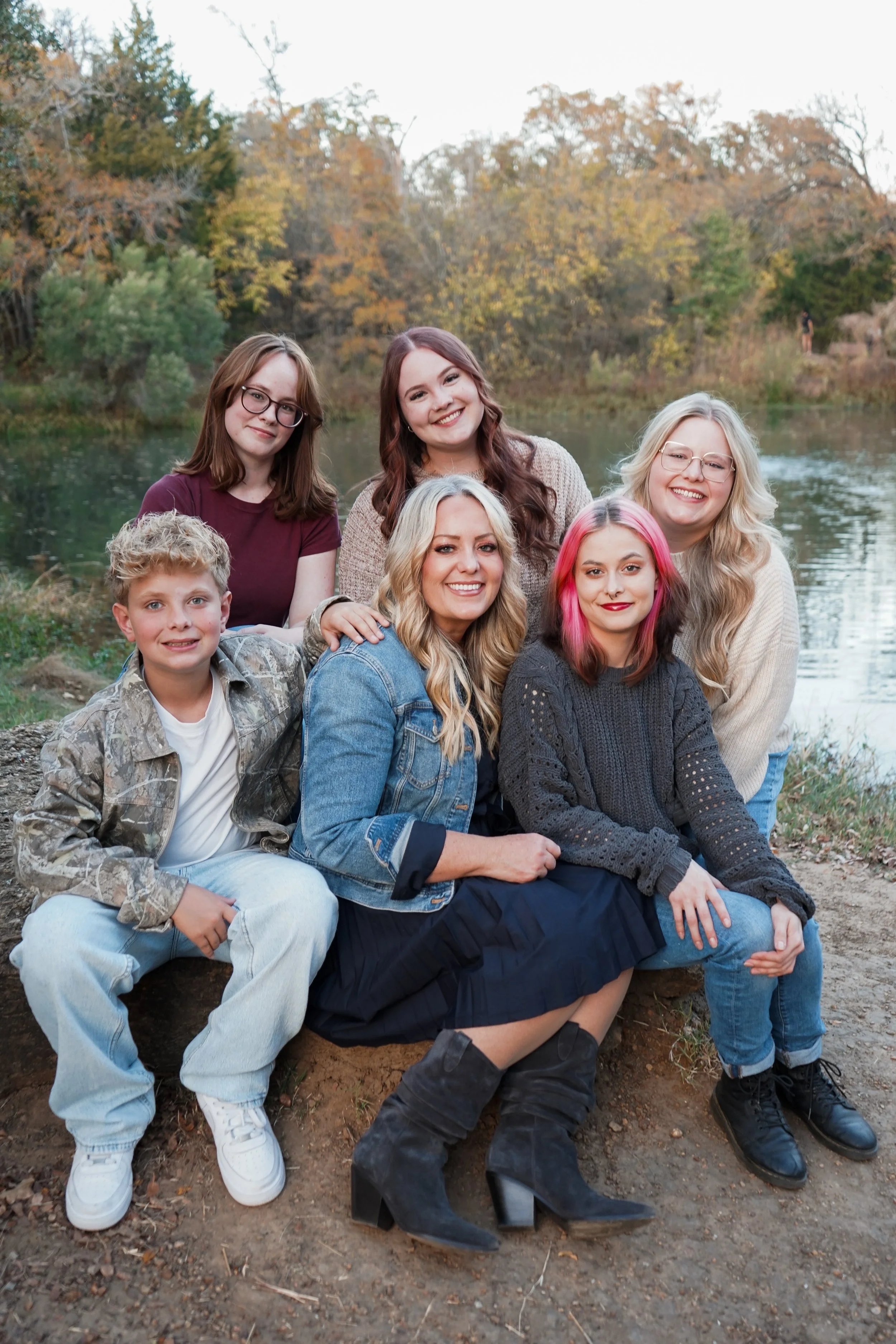 A group of seven people, including one young boy, six women, sitting and standing outdoors by a river during autumn, smiling at the camera.