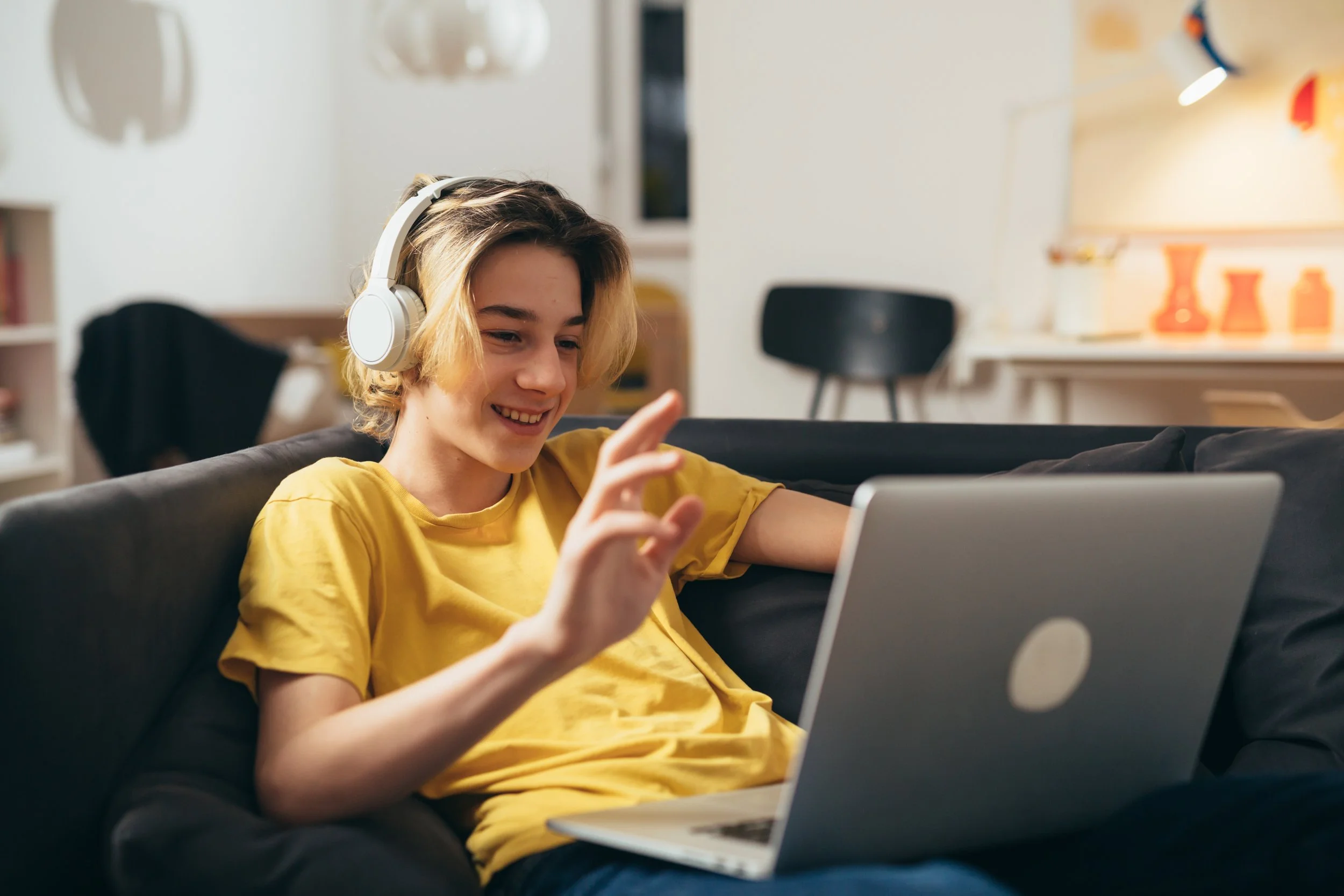 A young man with curly hair and glasses smiling as he looks at his laptop in a kitchen.