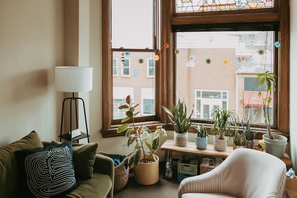 A corner of the office with large windows, plants on a shelf, a beige armchair and a green sofa.