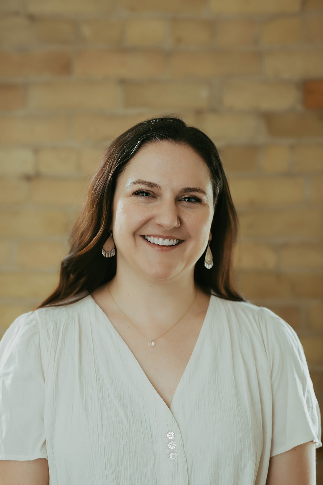 Smiling woman with long dark hair wearing earrings, a necklace, and a cream-colored blouse, standing in front of a brick wall.