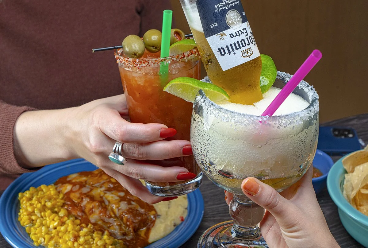 Two people are clinking glasses of cocktails, one with a lime wedge and salt on the rim and the other with green olives and a lime wedge. In the background, a plate of Mexican food with corn and sauce and a bowl of chips can be seen.