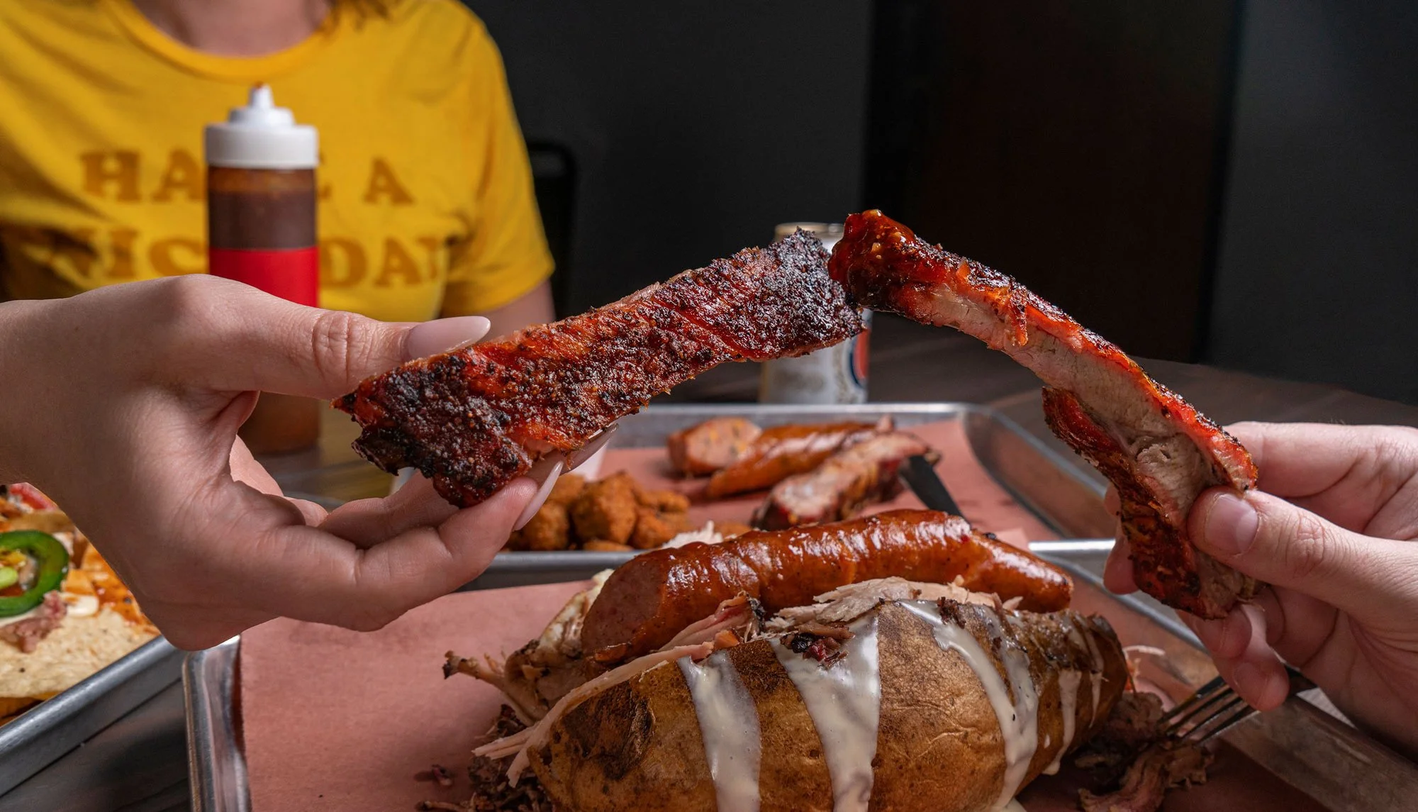 Two people are holding and sharing a rack of barbecue ribs above a tray of baked potato, sausage, and shredded pork; a woman in a yellow shirt is visible in the background.