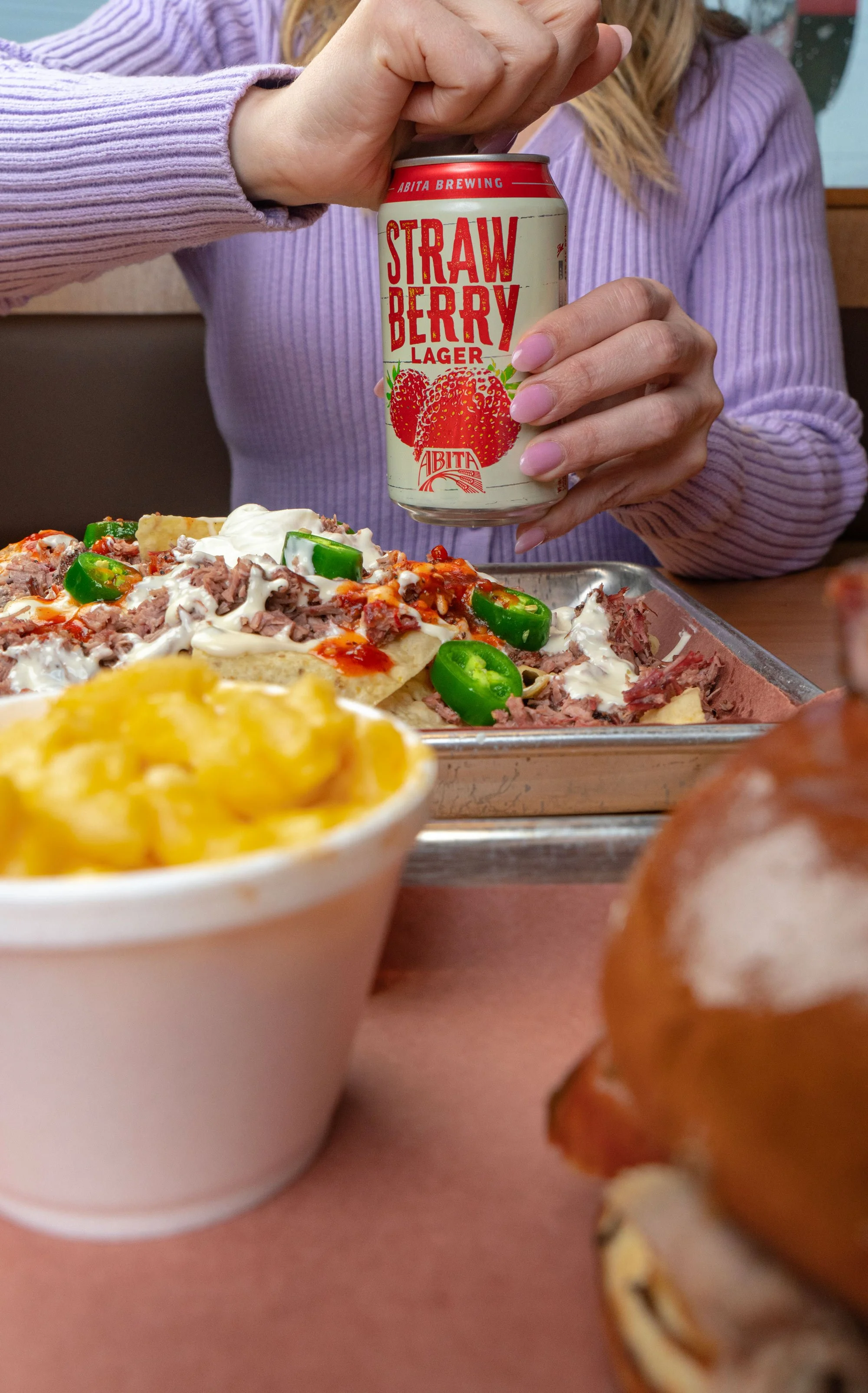 A person holding a can of strawberry lager beer next to a tray of nachos topped with jalapenos, sour cream, and meat, a bowl of macaroni and cheese, and a sandwich with a bun and filling.